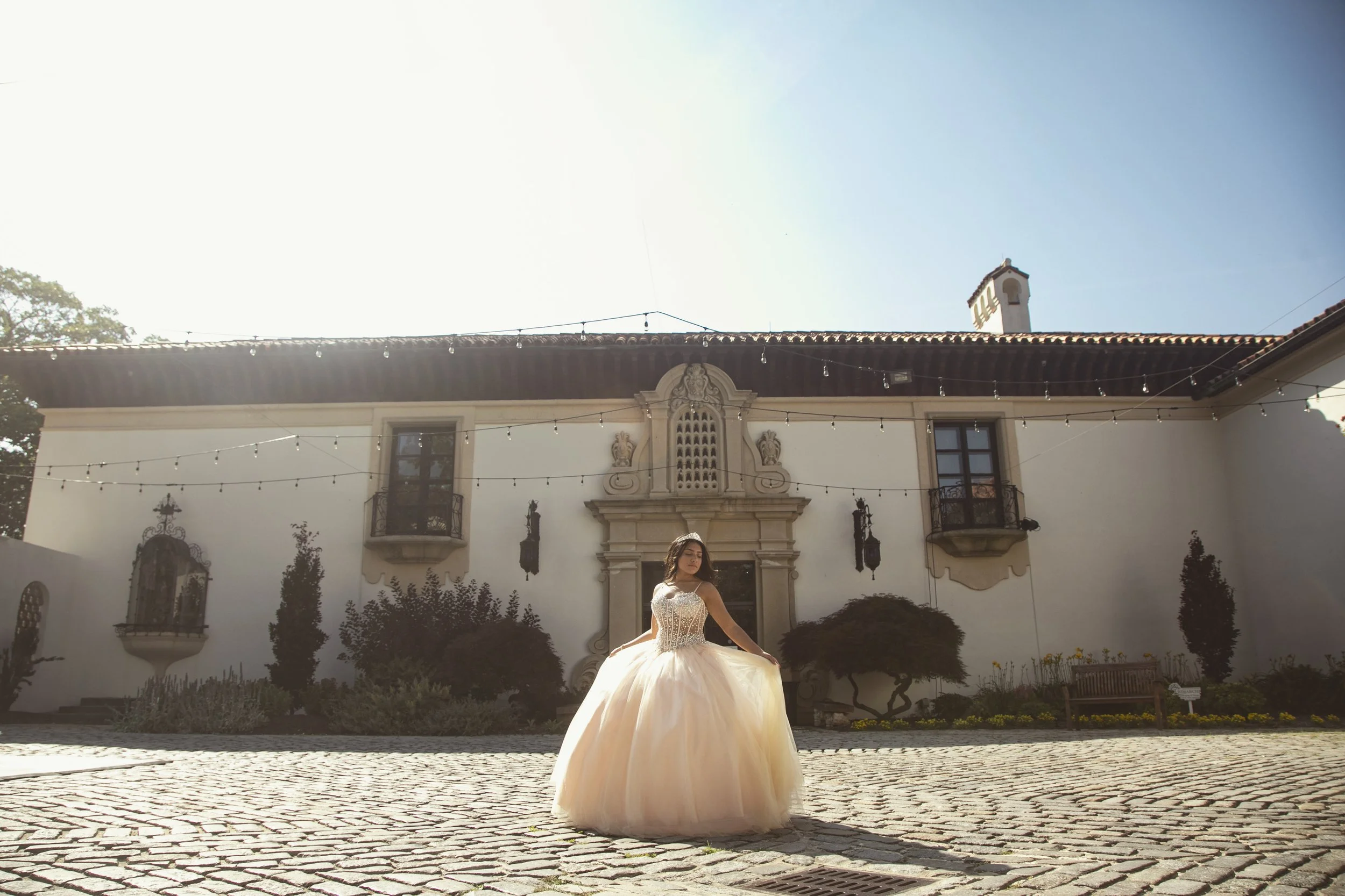 A woman in a wedding dress standing outside a large, elegant building with string lights hanging across the courtyard.