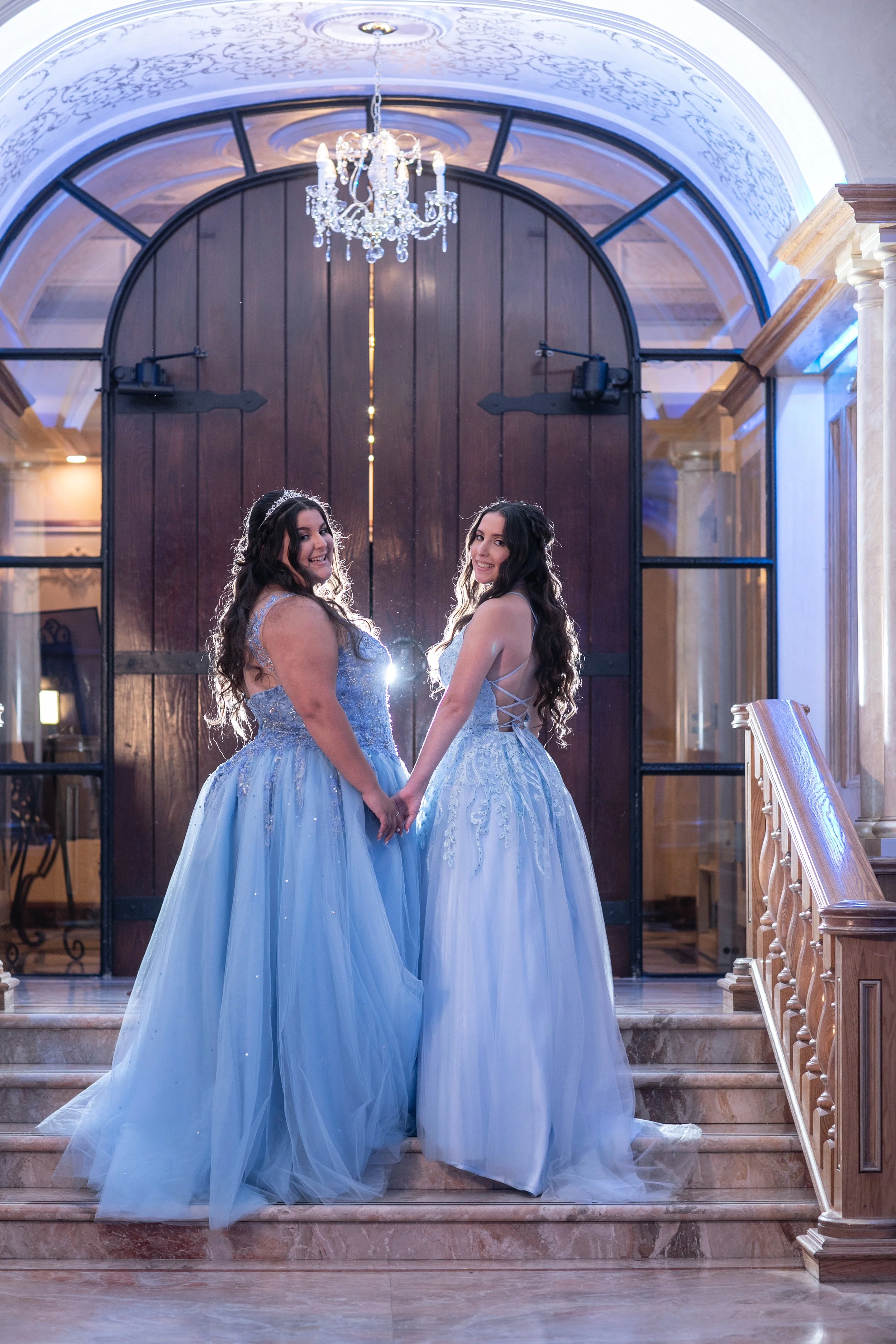 Two women in elegant blue ball gowns holding hands on a marble staircase in a grand hall with a chandelier and arched ceiling.