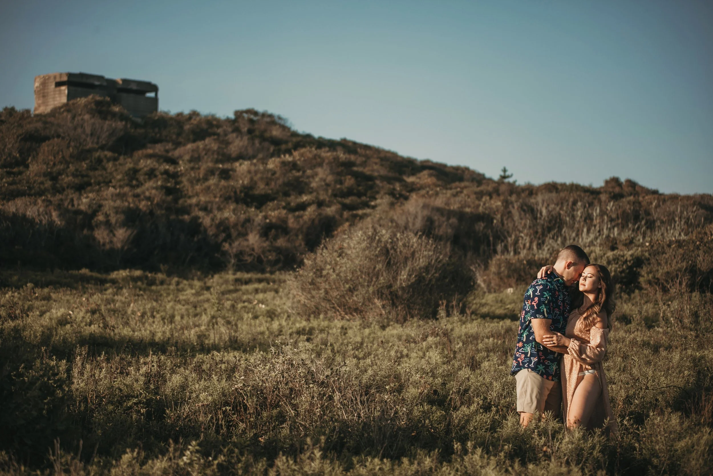 A couple embracing outdoors on a grassy hill with a forested landscape and a large structure on a hilltop in the background during late afternoon or early evening.