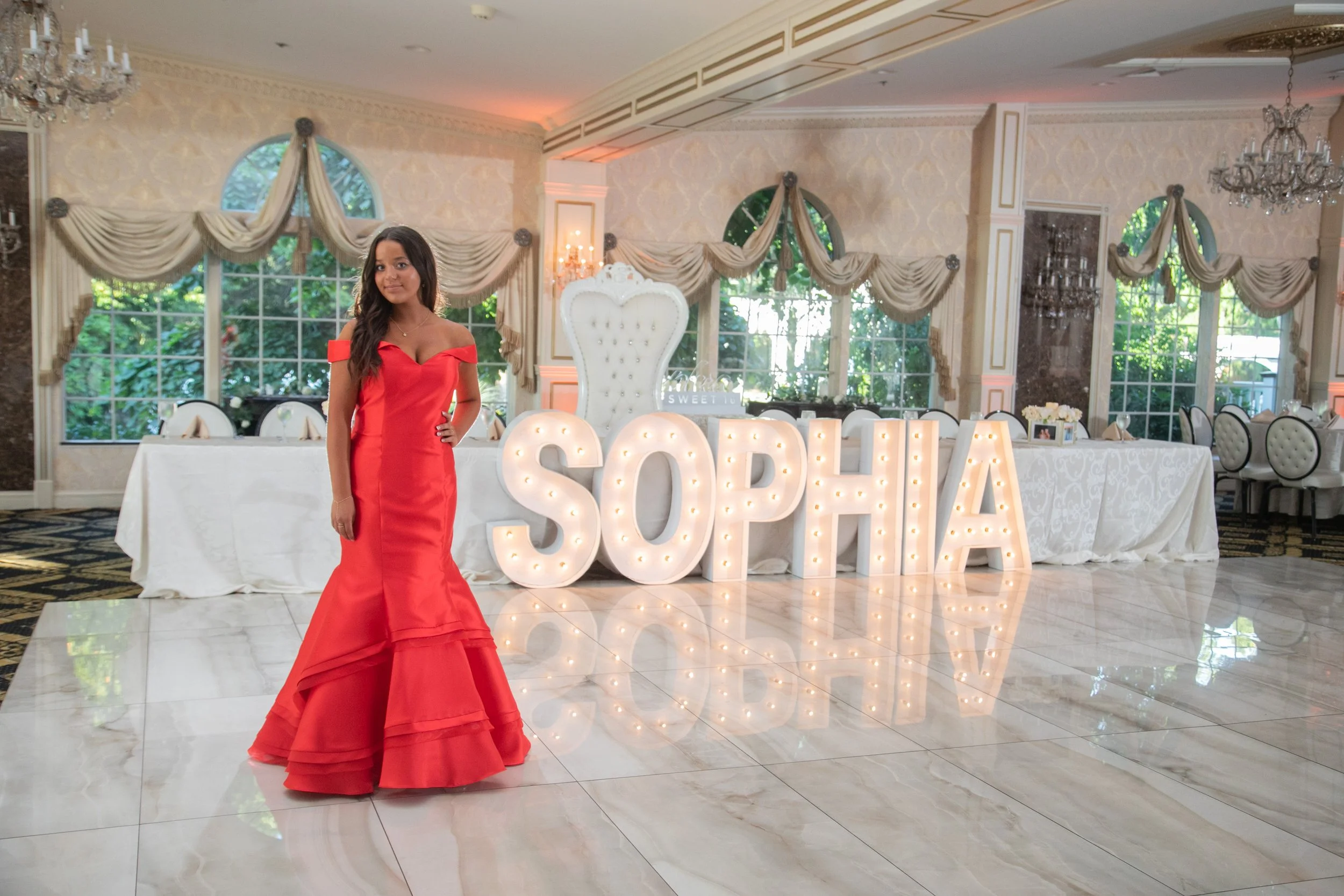 Young woman in a red formal dress standing in an elegant party hall with a large illuminated sign spelling SOFIA, decorated with cream-colored curtains, chandeliers, and greenery outside the windows.