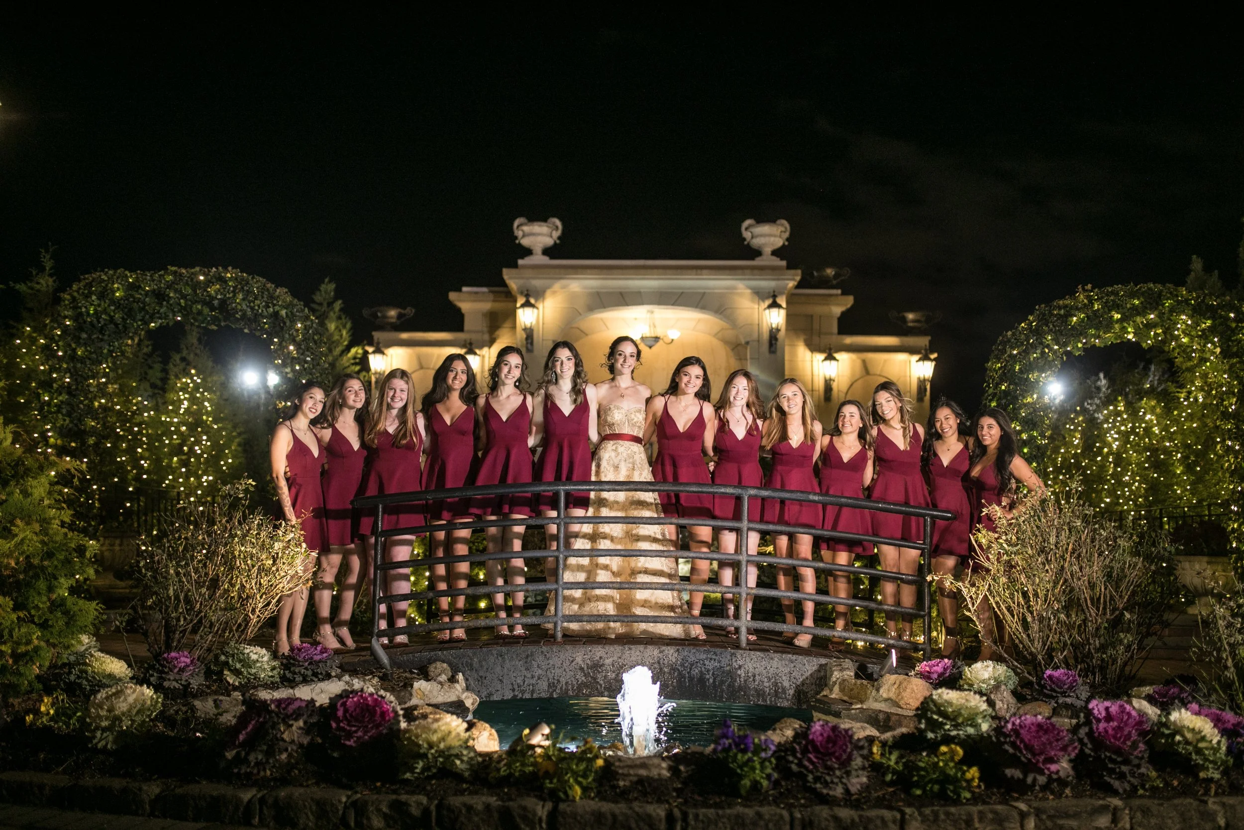 Group of young women in matching maroon dresses standing on a small bridge by a fountain at night, in front of a lit historic building with decorative urns on top and lush greenery with string lights.