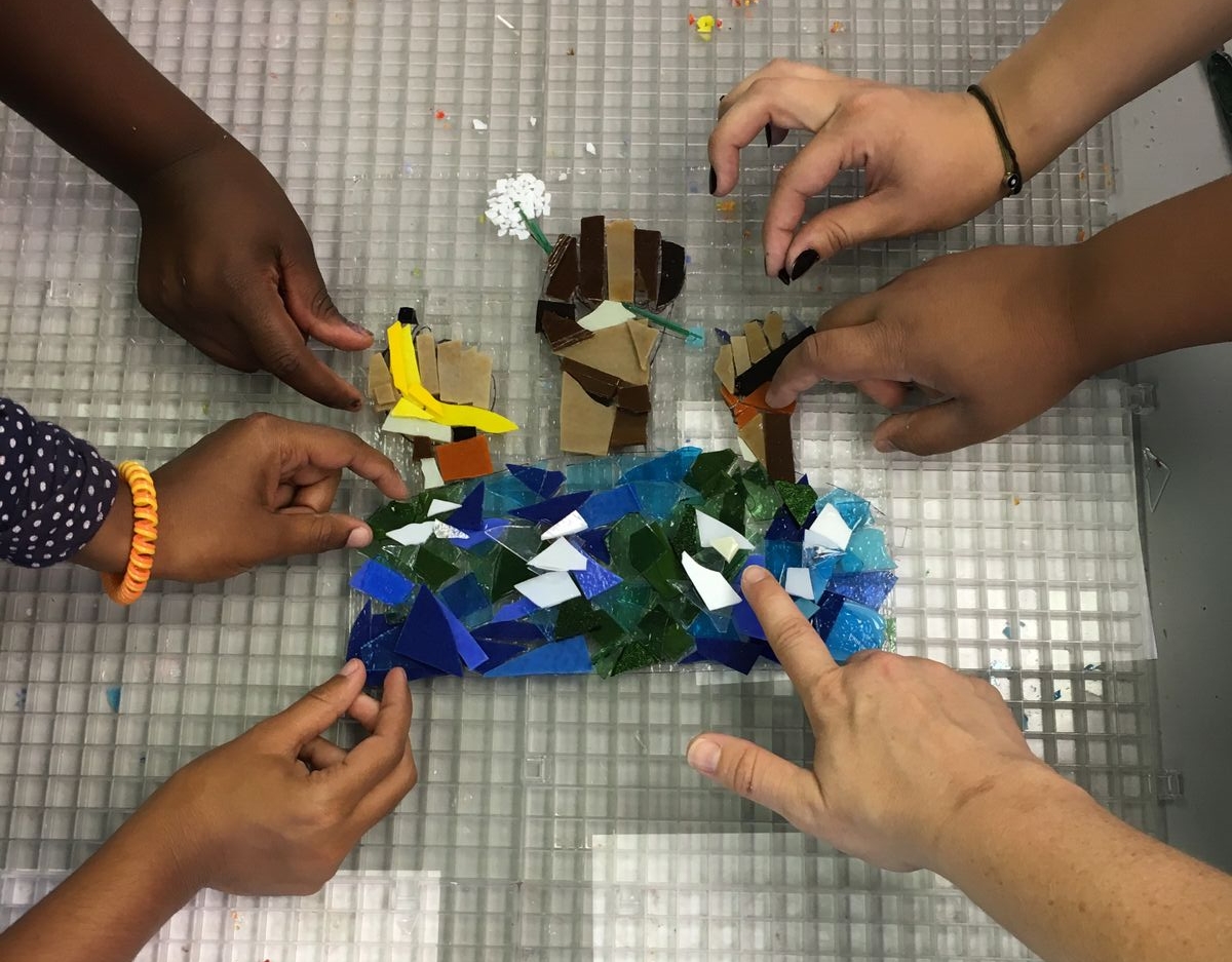 Students work on the CYC-Fellowship House mural together. The multicolored hands represents a diverse community and the blue-green circle at the bottom is the earth. The three hands are holding symbolic items: a banana (sharing food), a hammer (working together), and a flower (peace and beauty).