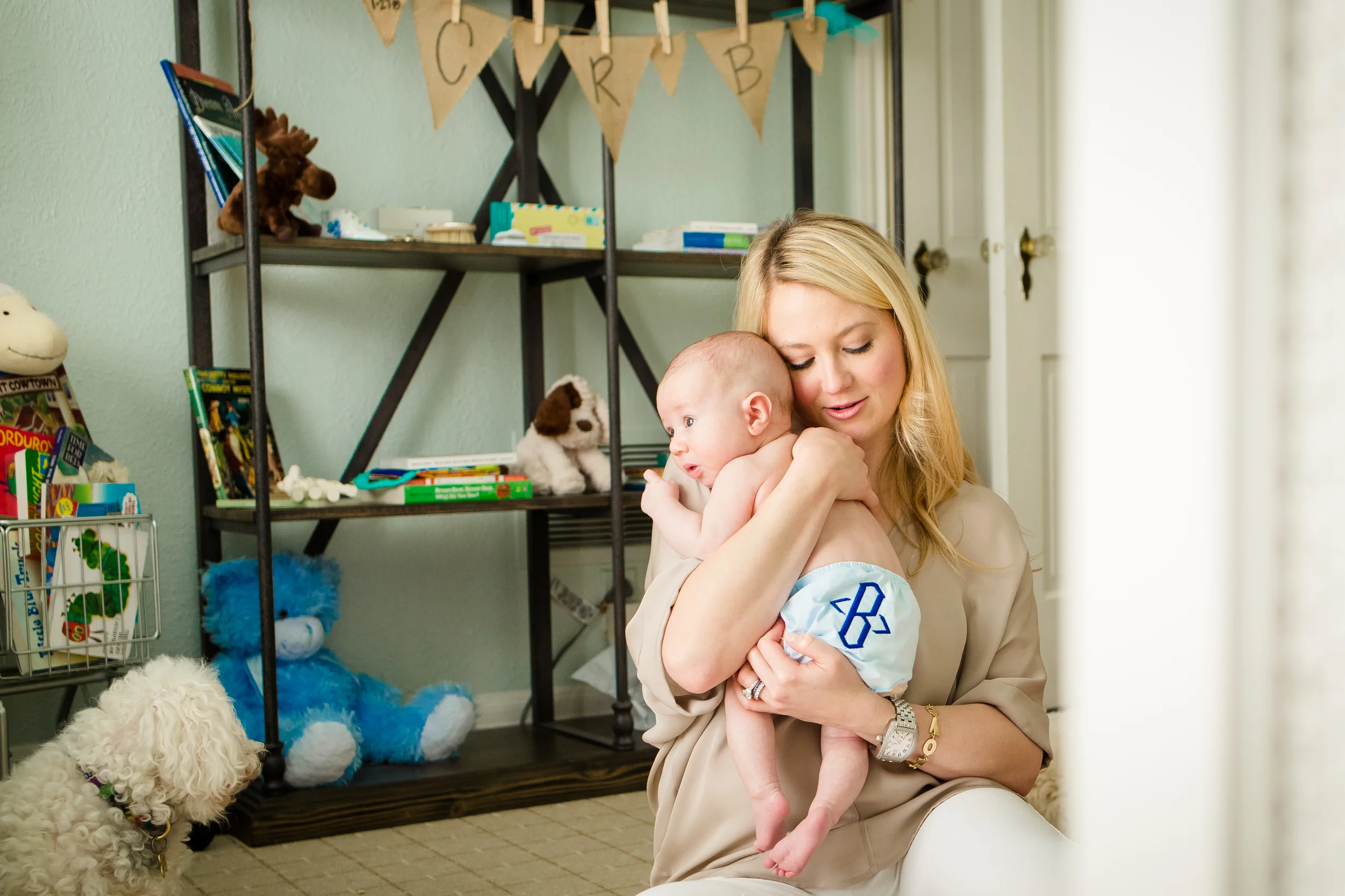 A woman with long blonde hair holding a baby in a room decorated for a birthday celebration. The woman is smiling with eyes closed, holding the baby close to her face. There are several stuffed animals and toys on a black metal shelf behind them, including a stuffed moose and a large blue fluffy creature. A dog with curly white fur is in the foreground. The room has soft green walls and a banner with the initials 'CRB' hanging above the shelf.