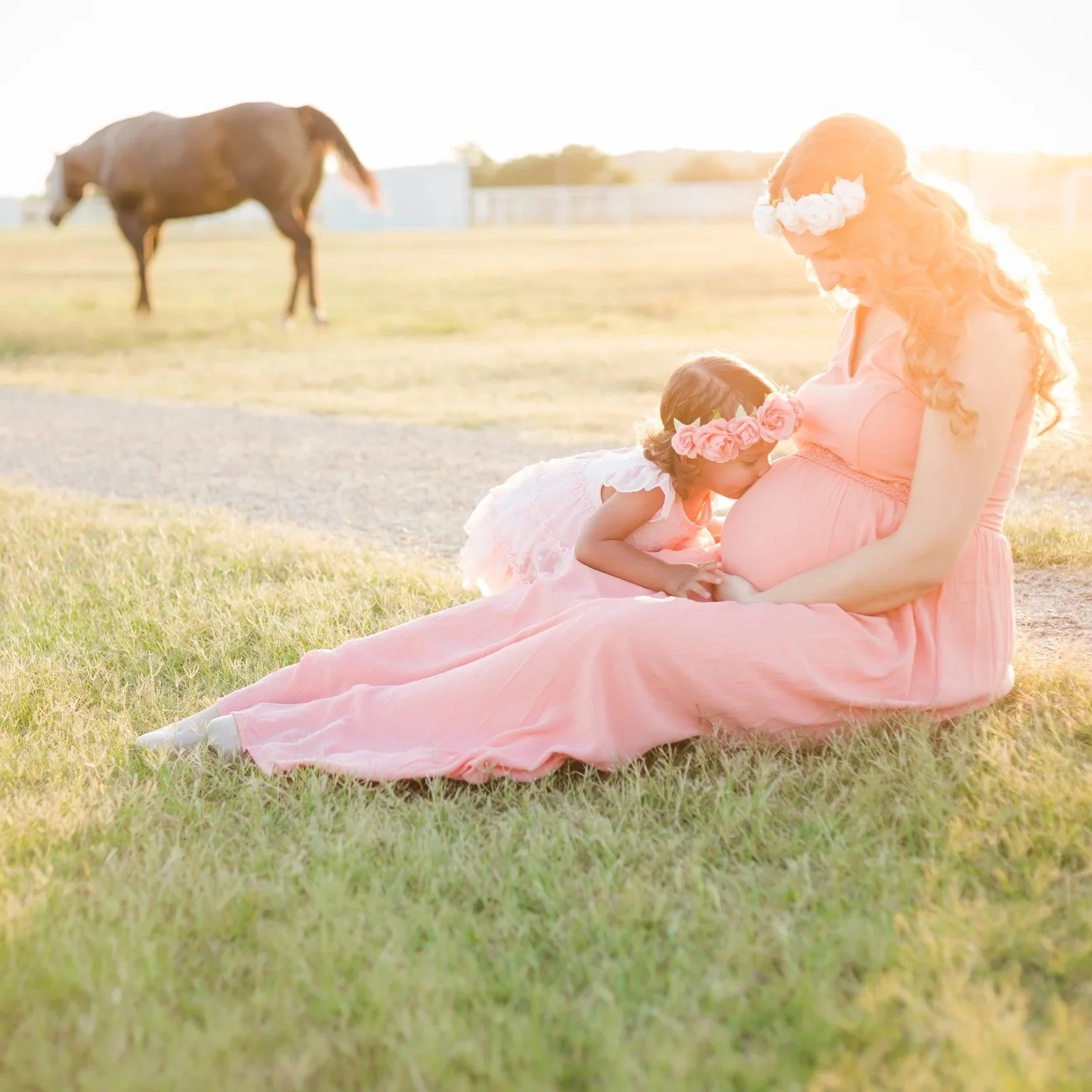 A pregnant woman in a pink dress and a girl with a flower crown sitting on grass at sunset.
