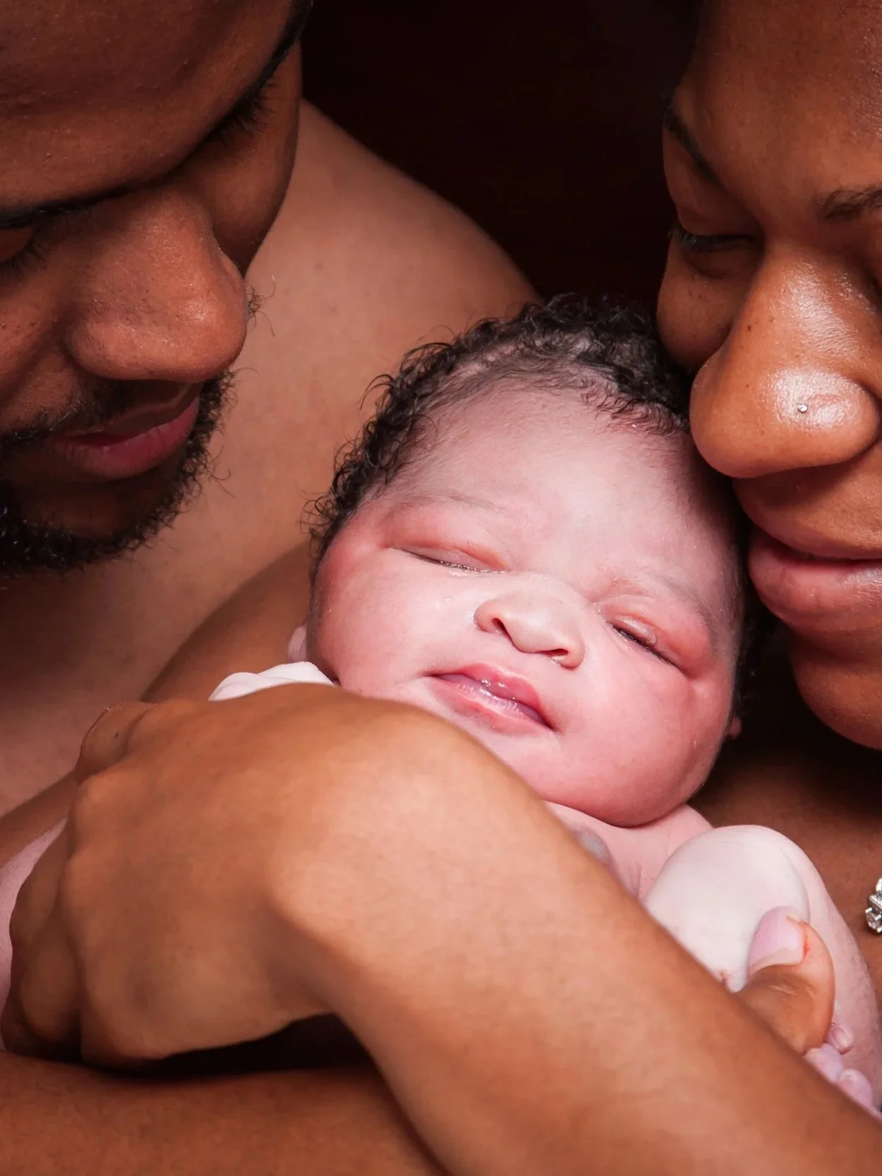 Close-up of a newborn baby being held by two adults, with the adults's faces close to the baby's face, in an intimate moment of bonding.
