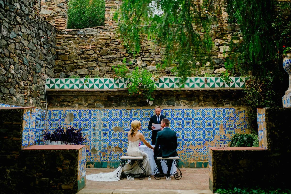 Wedding ceremony at Torre Bellesguard in Barcelona, bride and groom seated in ornate wrought iron chairs against Gaudí's iconic mosaic tile wall