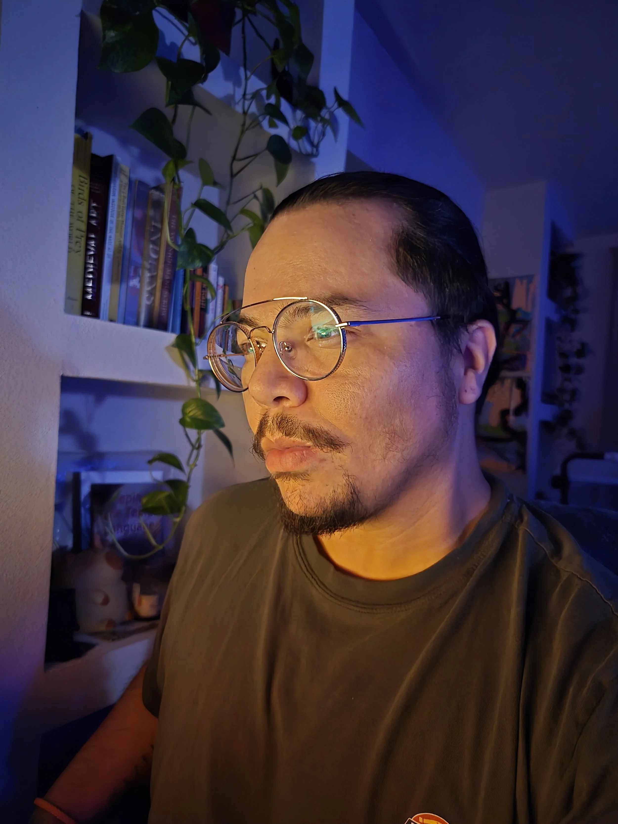 Close-up of a man with glasses, dark hair, and facial hair, looking down and to the side, in a room with blue lighting, a bookshelf with books, and a green plant in the background.