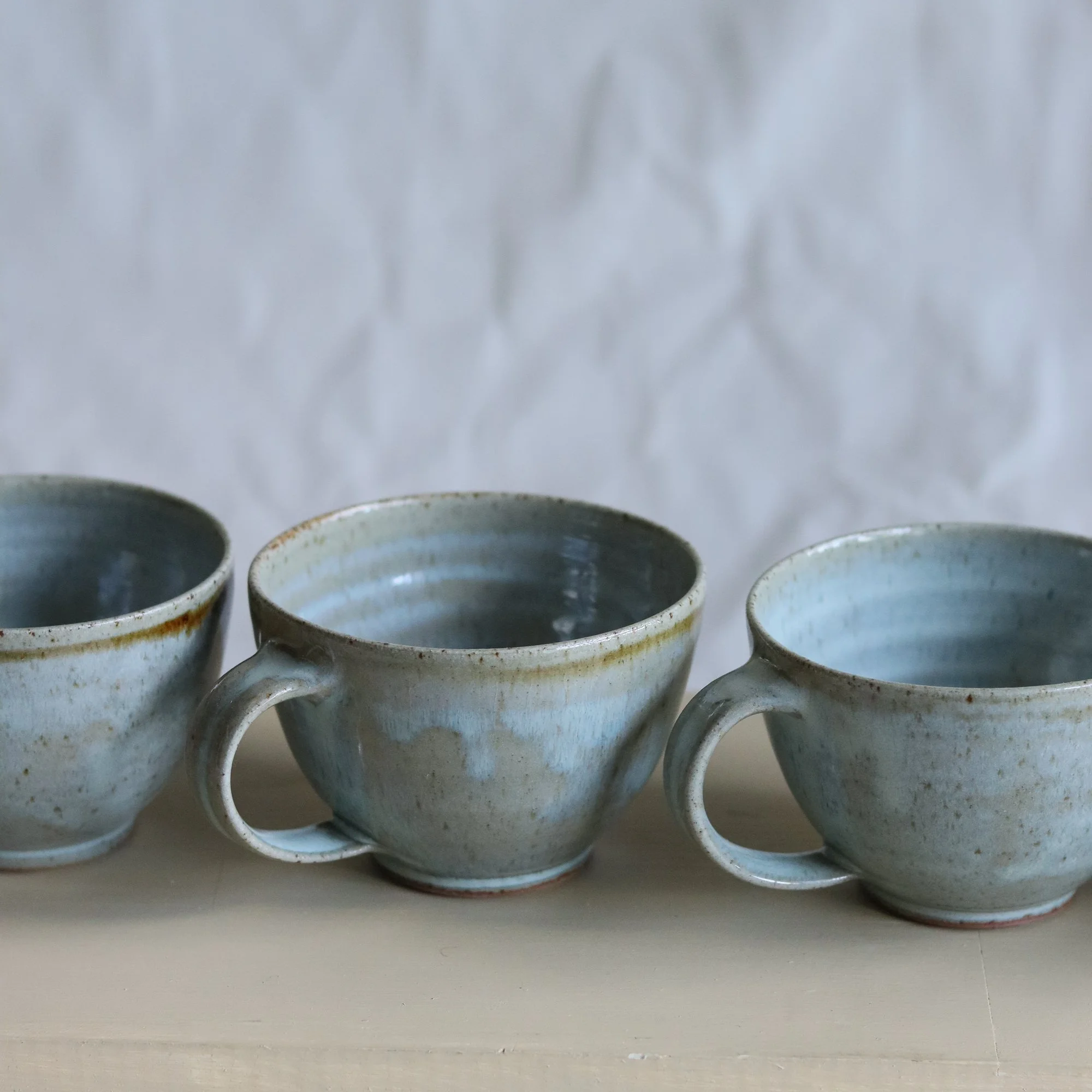 Row of curved mugs in pale blue chun by Pottery West