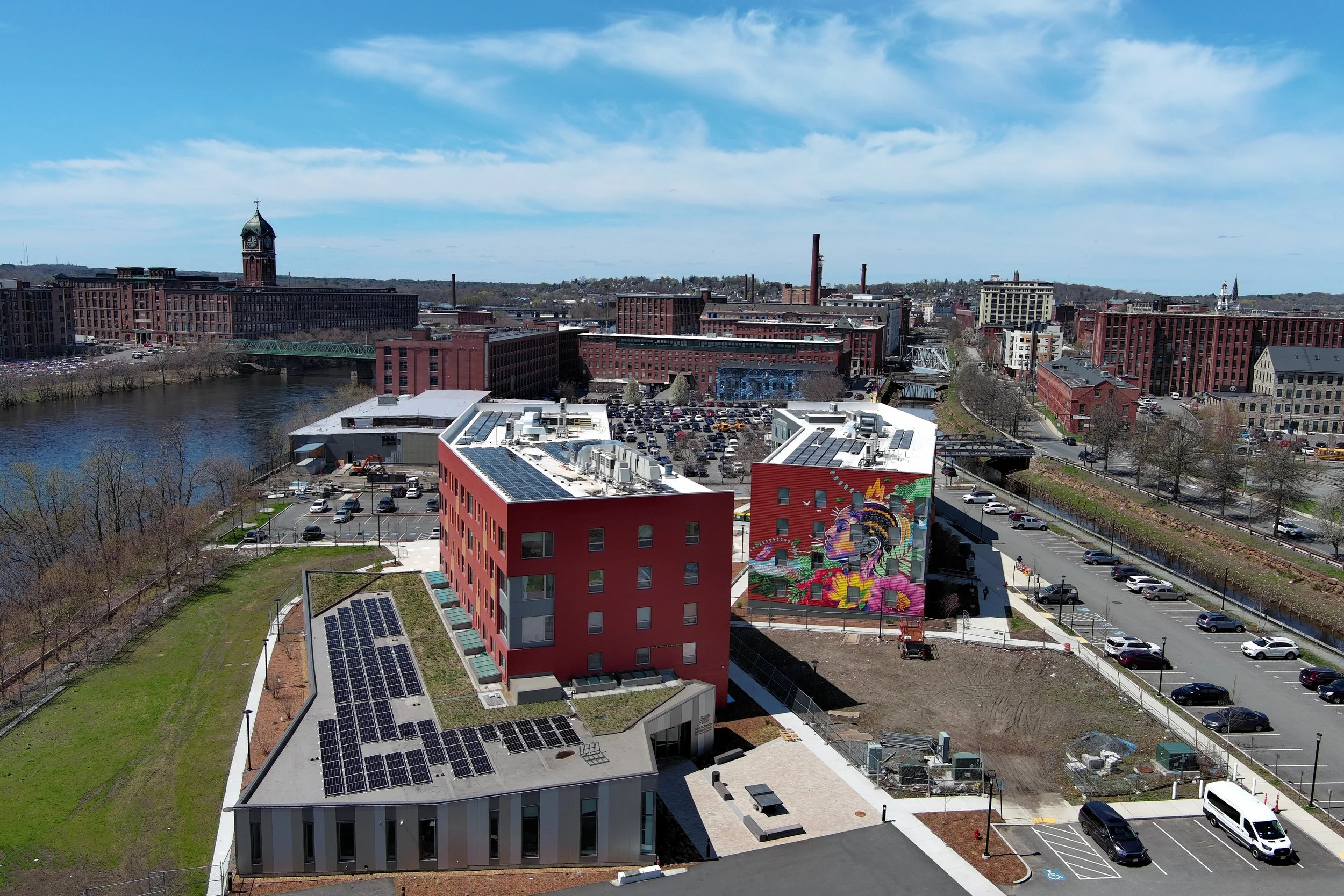 A cityscape with a river in the foreground, modern buildings including one with solar panels, a colorful mural on a red building, parking lots, and a downtown skyline with a clock tower, factories, and church steezes under a partly cloudy sky.