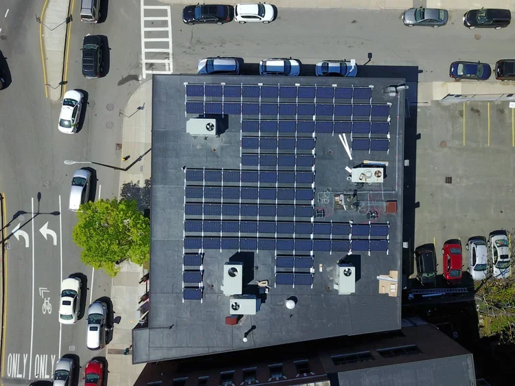 A rooftop with solar panels installed on it, surrounded by a parking lot with various cars, and a small green tree beside the building.