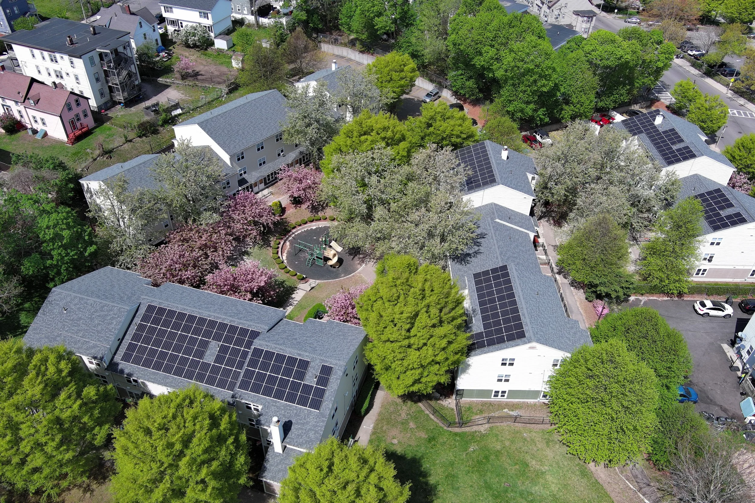 Aerial view of a neighborhood with houses, trees, and some with solar panels on the roofs, along with parking areas and a playground.