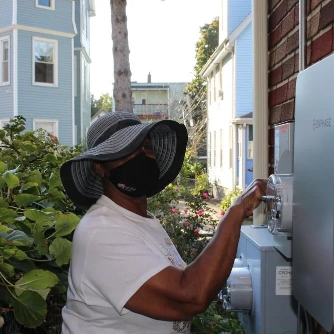 A woman wearing a large brim hat and face mask using a coin-operated laundry machine outdoors in a residential area with colorful houses and greenery.