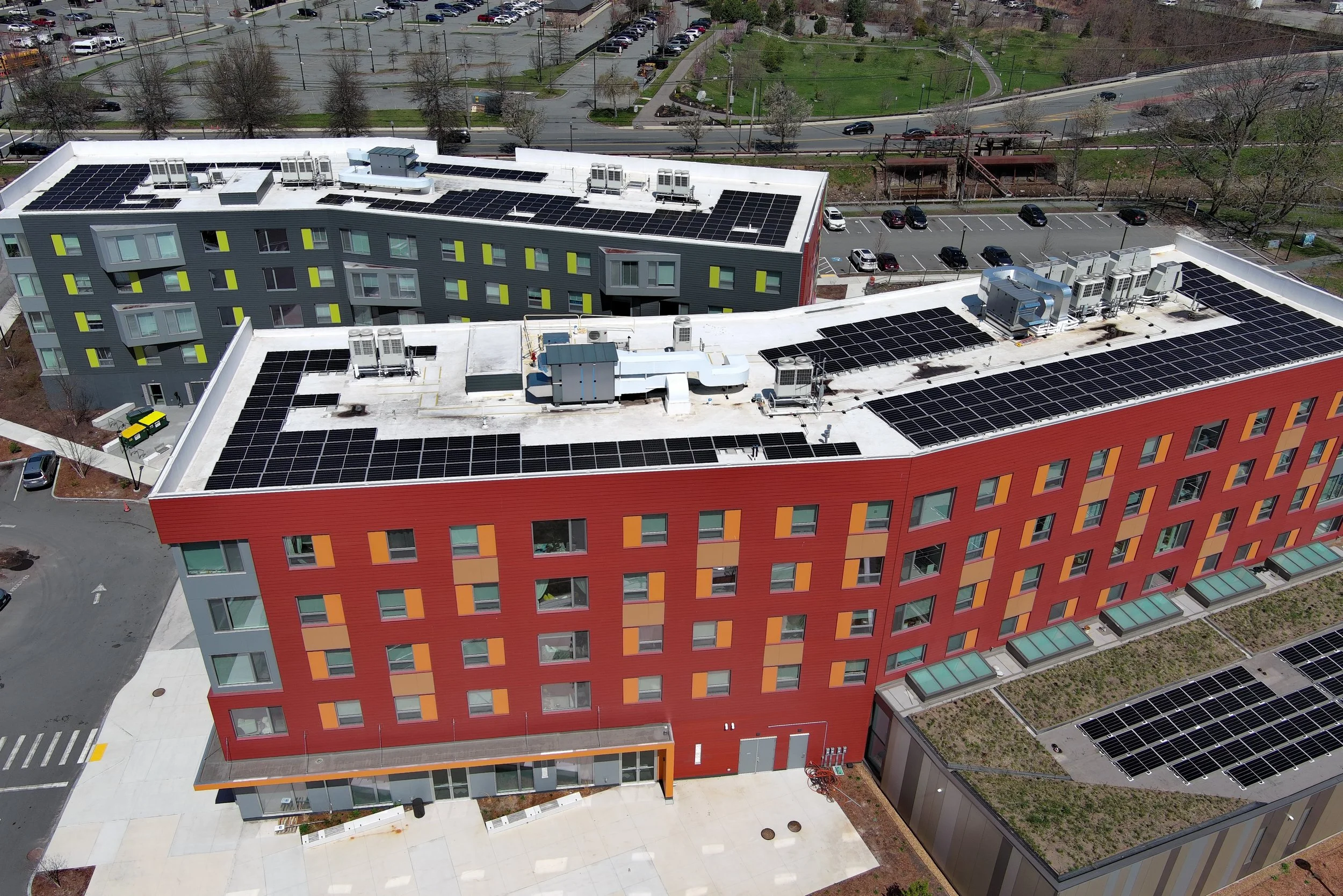 Aerial view of a modern multi-story building with solar panels on the roof, green spaces surrounding it, parking lots, and nearby roads with cars.