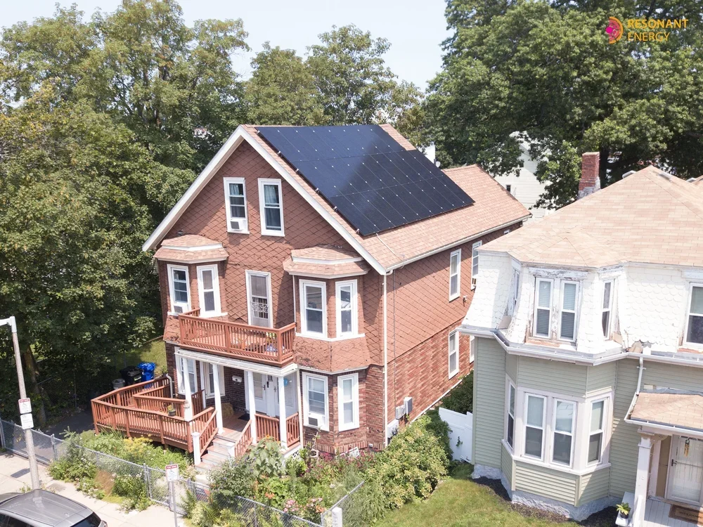 A two-story brick house with a deck and solar panels on the roof, surrounded by trees and neighboring houses.