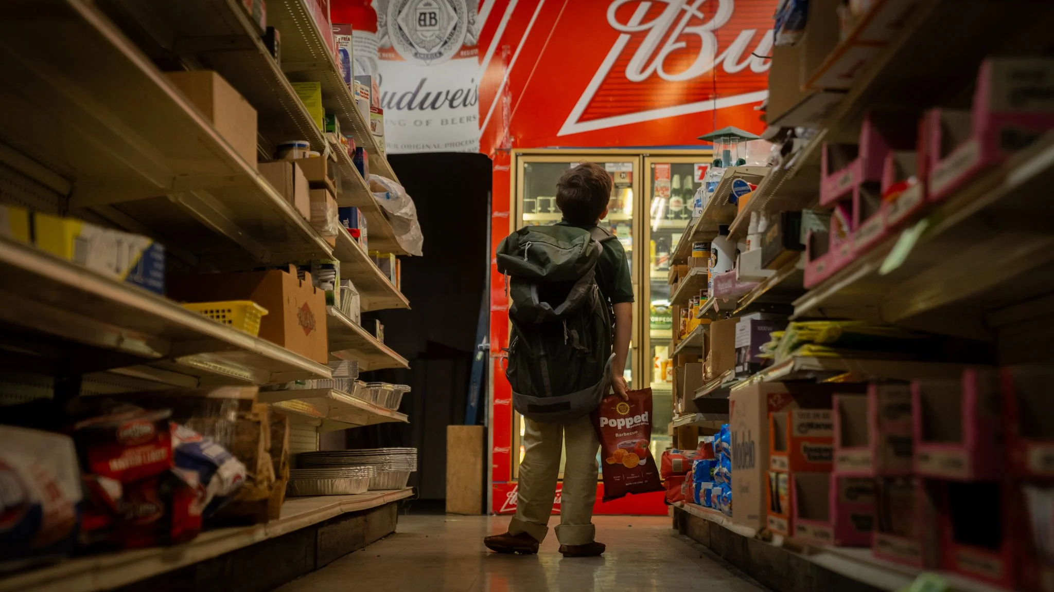 A young boy with a backpack at a convenience store aisle, facing a cooler with beverages, and holding a bag of potato chips.