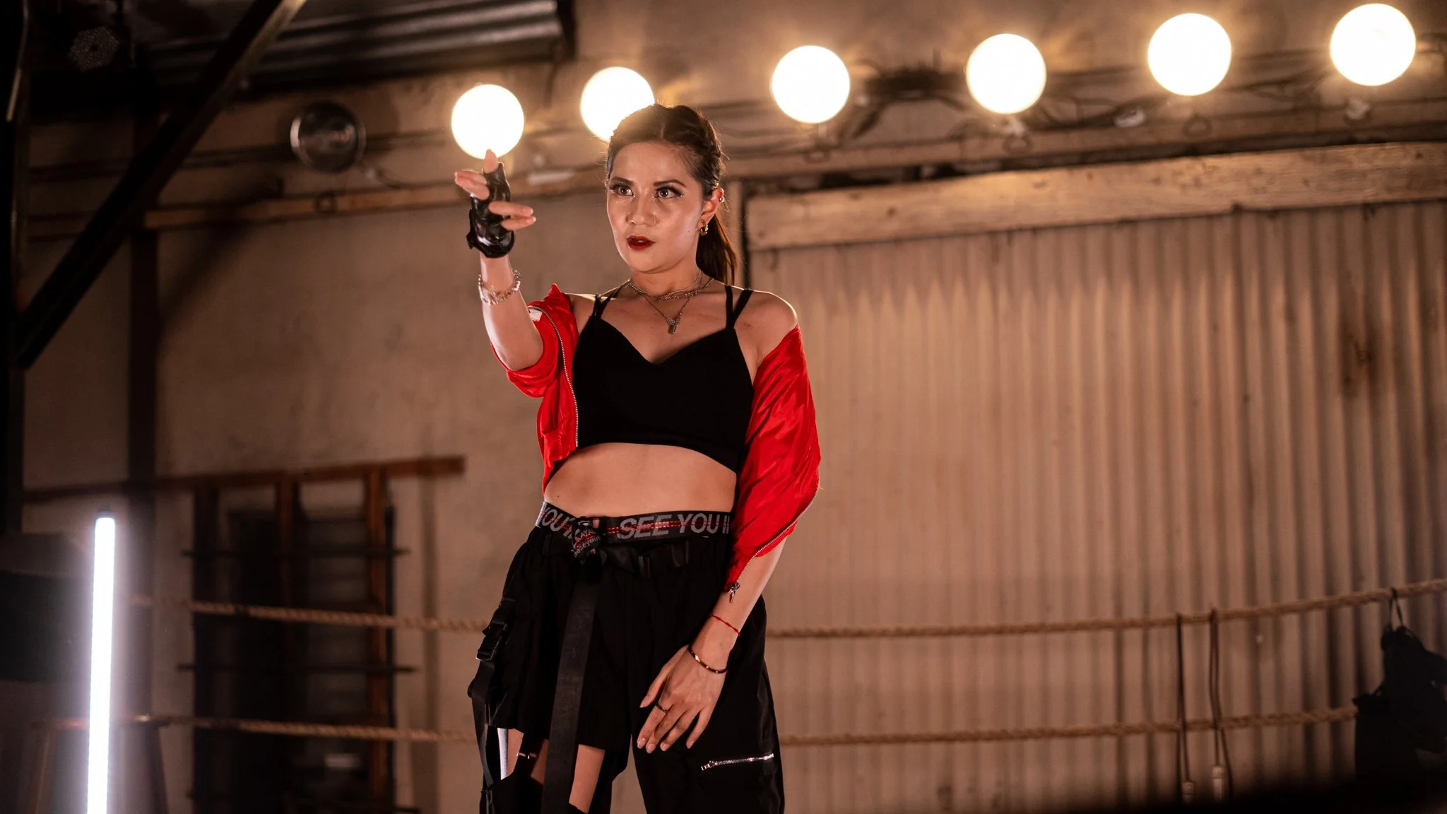 A young woman in a gym or dance studio pointing towards the camera with one hand, wearing a black crop top, red jacket, black pants, and gloves, with a confident expression.