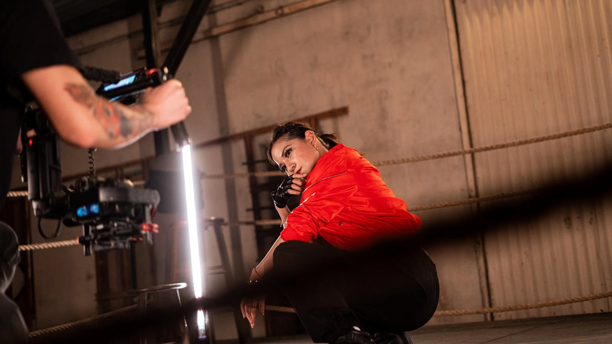 A woman wearing a red jacket and black gloves is sitting against a wall in an industrial-style room, posing for a photo being taken with professional camera equipment.