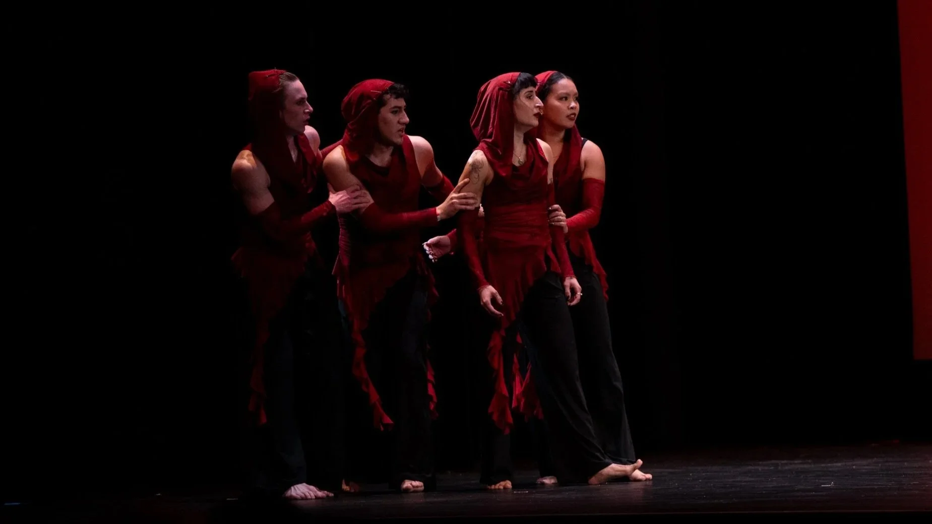 Four performers dressed in red and black costumes are standing in a line on a dark stage, facing to the right. They have serious expressions, and the lighting highlights their faces and outfits against the black background.
