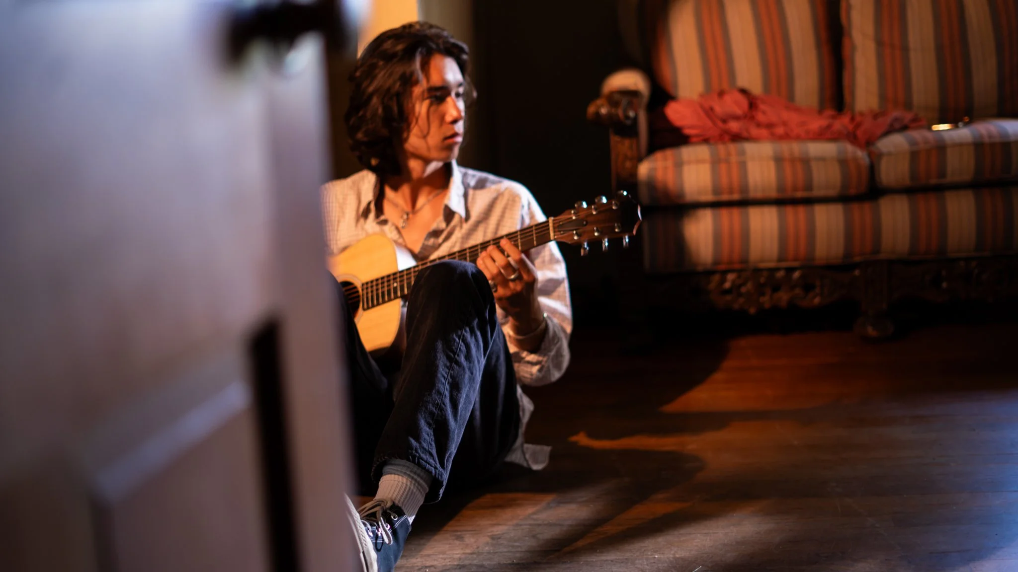 Young person with long hair sitting on the floor playing an acoustic guitar in a dimly lit room, with a striped sofa and patterned curtain in the background.
