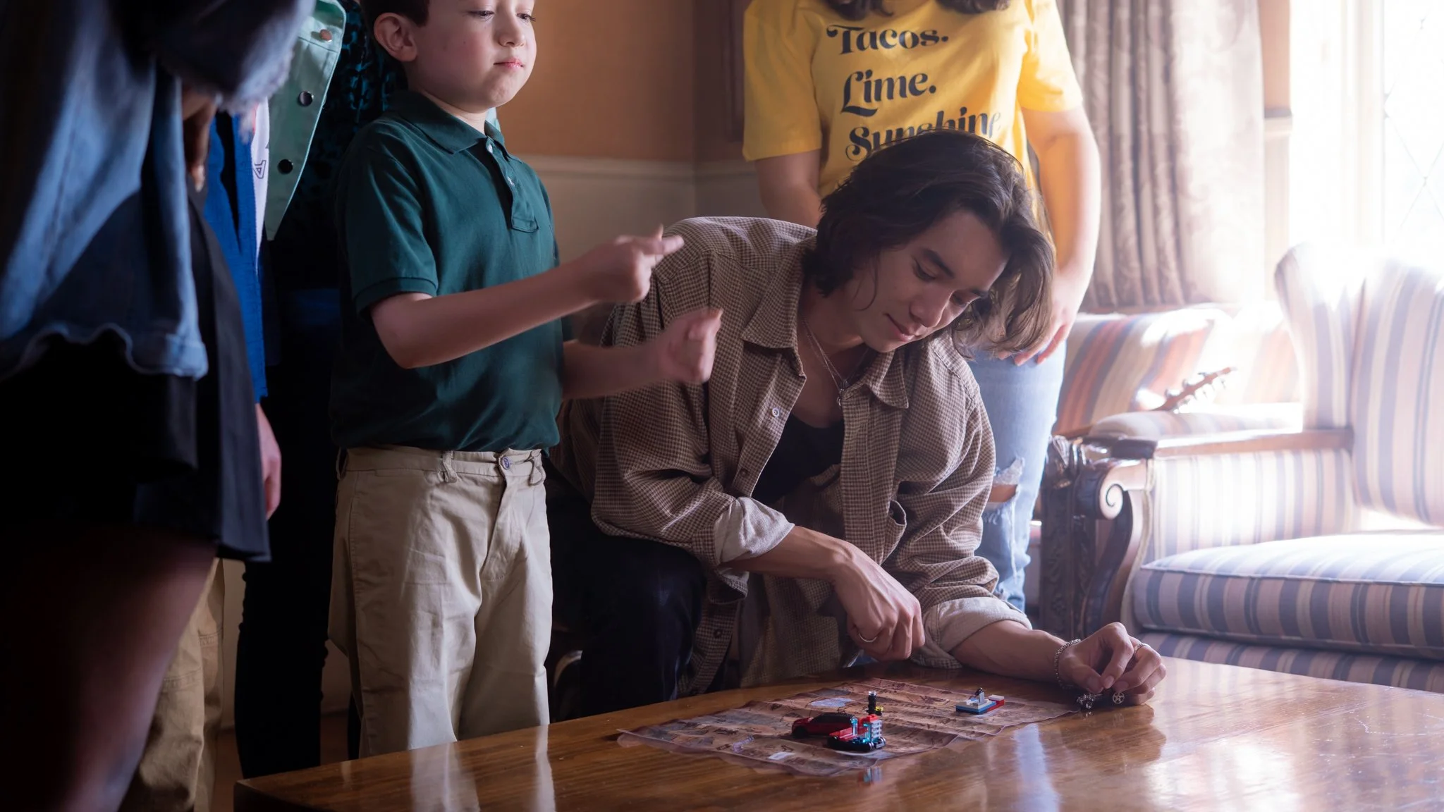A group of four people gathered around a table playing with small toys or game pieces. One person is sitting, leaning over the table, while the others stand close by. The room is warmly lit with sunlight coming through a window.