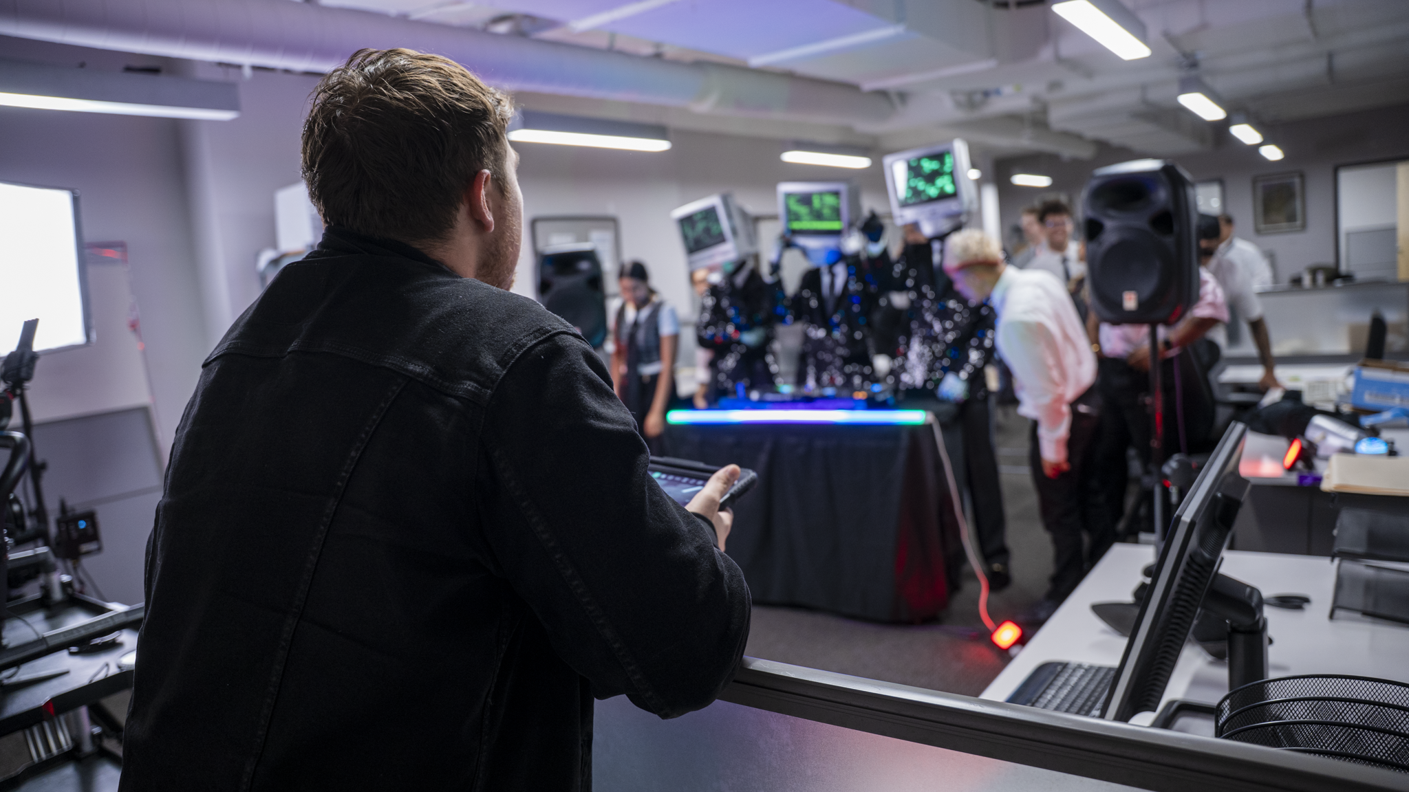 A young man with brown hair, wearing a black denim jacket, using a smartphone in a busy tech conference or event with several monitors displaying green and purple graphics in the background.