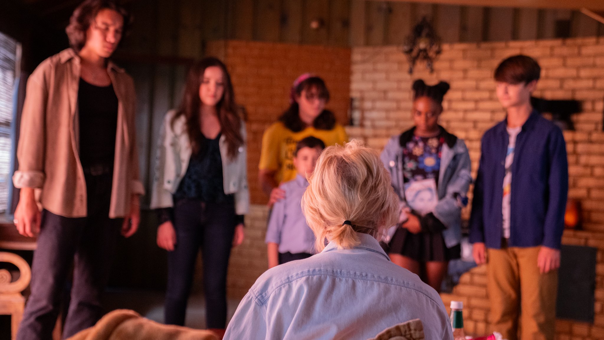 Group of people, including children and adults, standing together in a room with brick walls, facing a person with blonde hair tied back, who is seated with a light-colored shirt.
