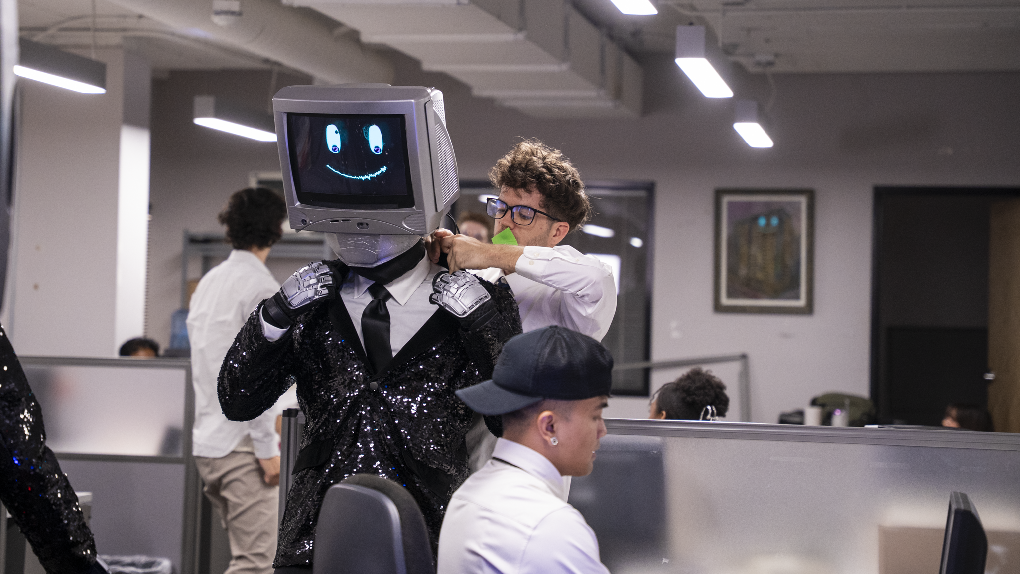 A person with a computer monitor head displaying a smiling face with long blue eyes in an office environment.