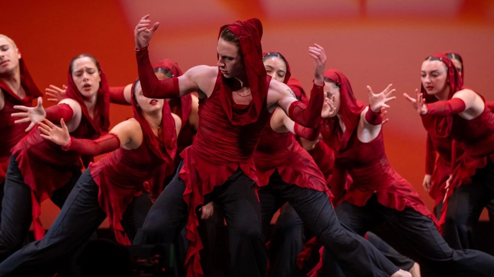 Group of dancers in red costumes performing on stage with an orange background.