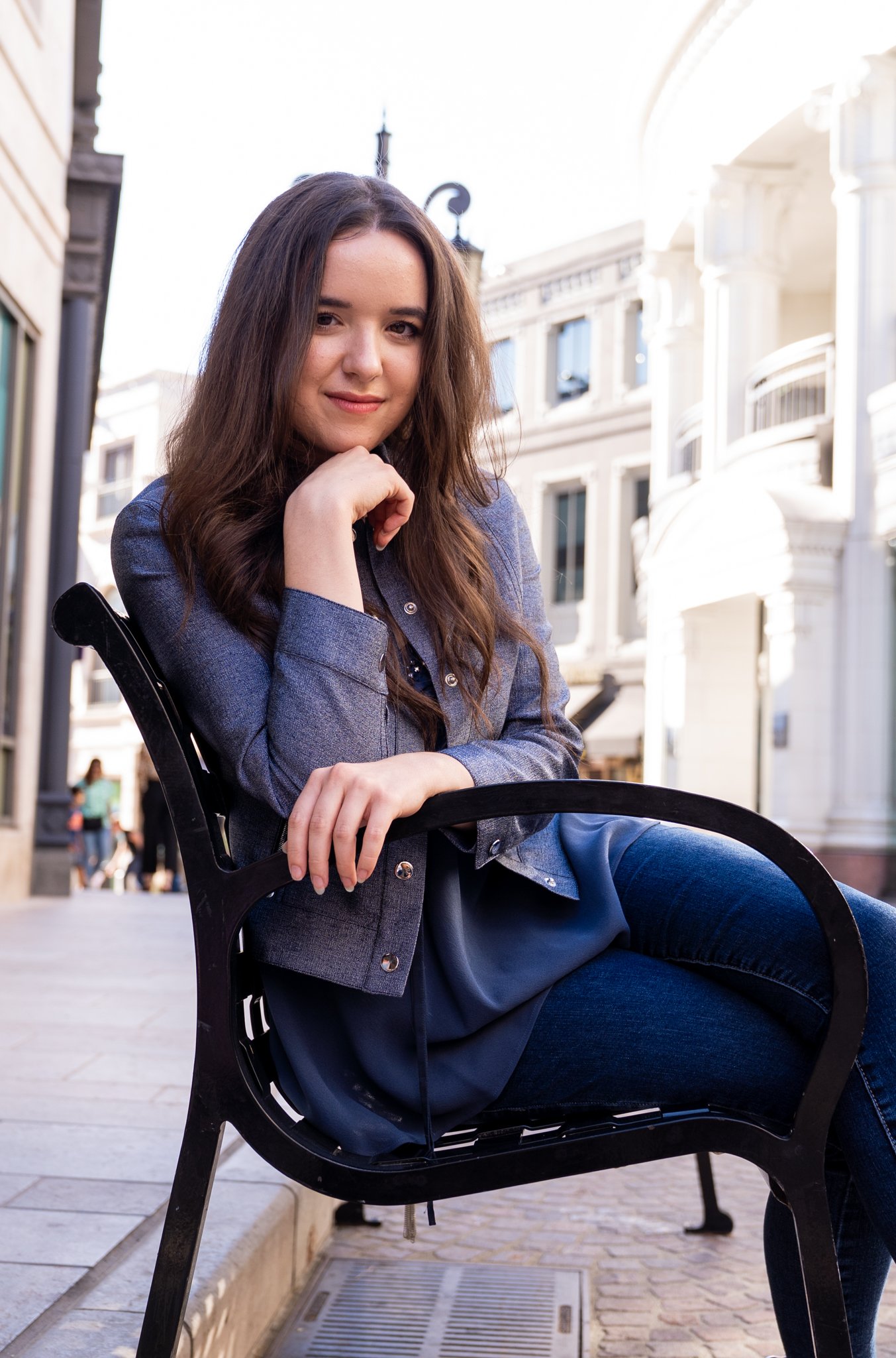 A young woman with long brown hair sitting on a black bench outdoors on a city street.