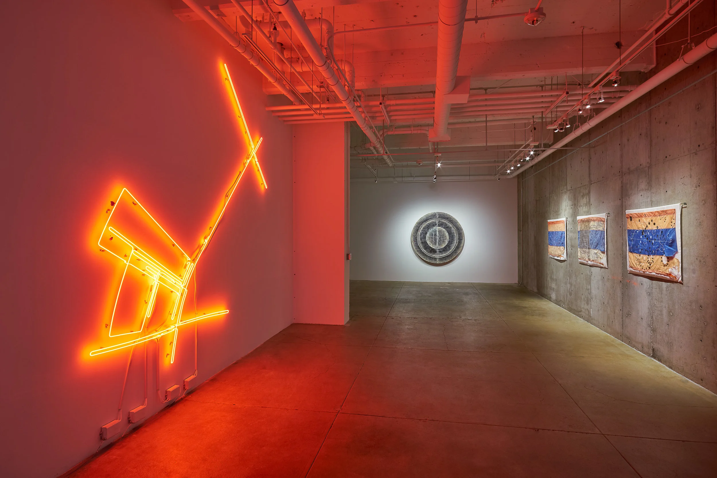 Exhibition documentation of Omar Mismar The Path of Love #03 , 2013-2014, neon, 126 x 126 inches (left), Taraneh Hemami Recounting , 2011, pigment and inkjet prints on paper and vellum, wax, wood panels, 66 inches diameter, 7 inches deep (center) and Guillermo Galindo Siguiendo Los Pasos del Niño Perdido, 20 Miles East of Laredo , and Cartografía del Espiritu/Cartography of the Spirit, all 2017, acrylic on beacon flags used by humanitarian aid group Water Stations (right). Image courtesy of the artists and Gallery Wendi Norris, San Francisco.