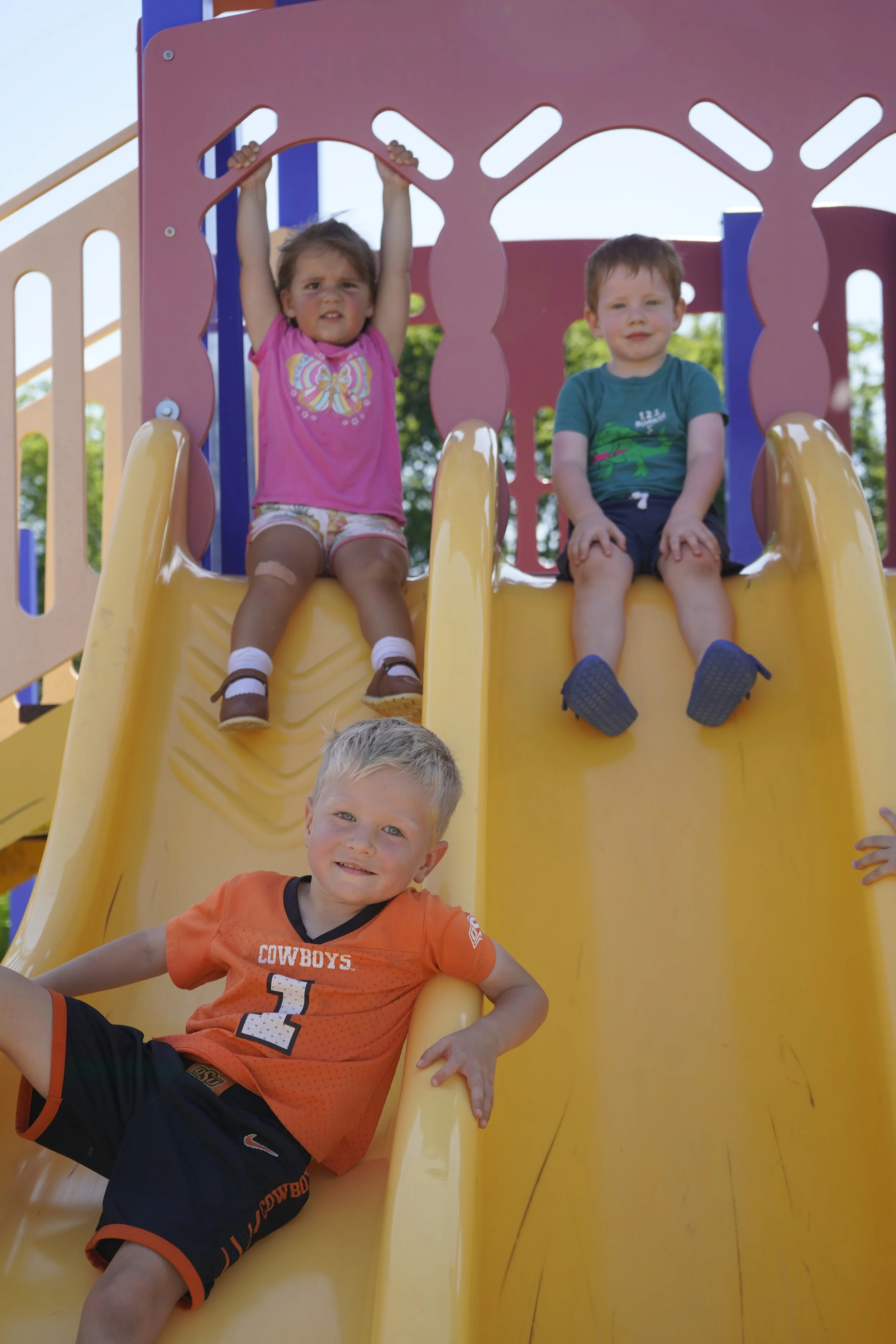 Three children playing on a yellow slide at a playground. The girl in pink is hanging from the top of the slide, the girl in green and black is sitting on the slide near the top, and the boy in orange is sliding down at the bottom. The background shows a blue sky and some trees.