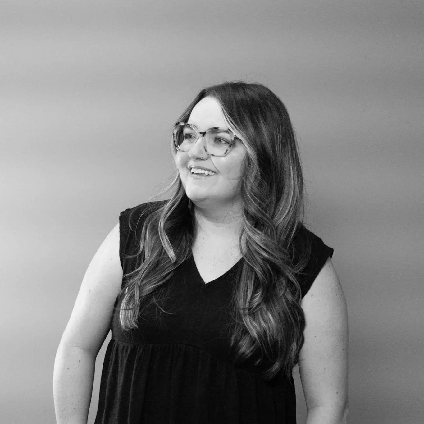 Black and white photo of a woman with long wavy hair, glasses, and a sleeveless black dress, smiling and looking to the side, standing against a plain background.