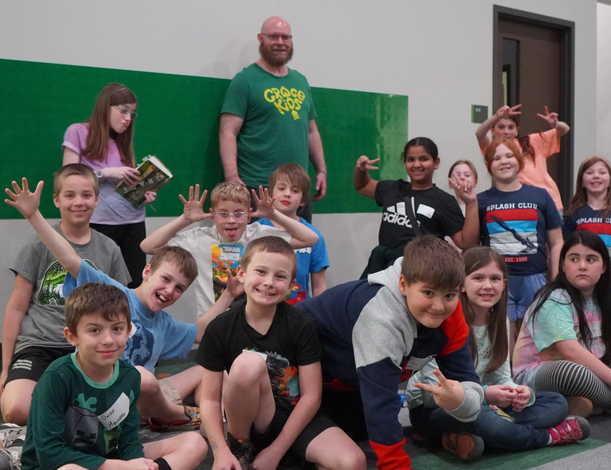A group of children and one adult man are gathered in a room, posing for a group photo. The children are smiling and making playful gestures. The man is standing in the middle, smiling, wearing glasses, a green t-shirt with yellow text that says 'Grace Kids,' and has a beard. The children are dressed casually, some with name tags, and are sitting or kneeling on the floor in front of a wall with a green section and a door to the right.