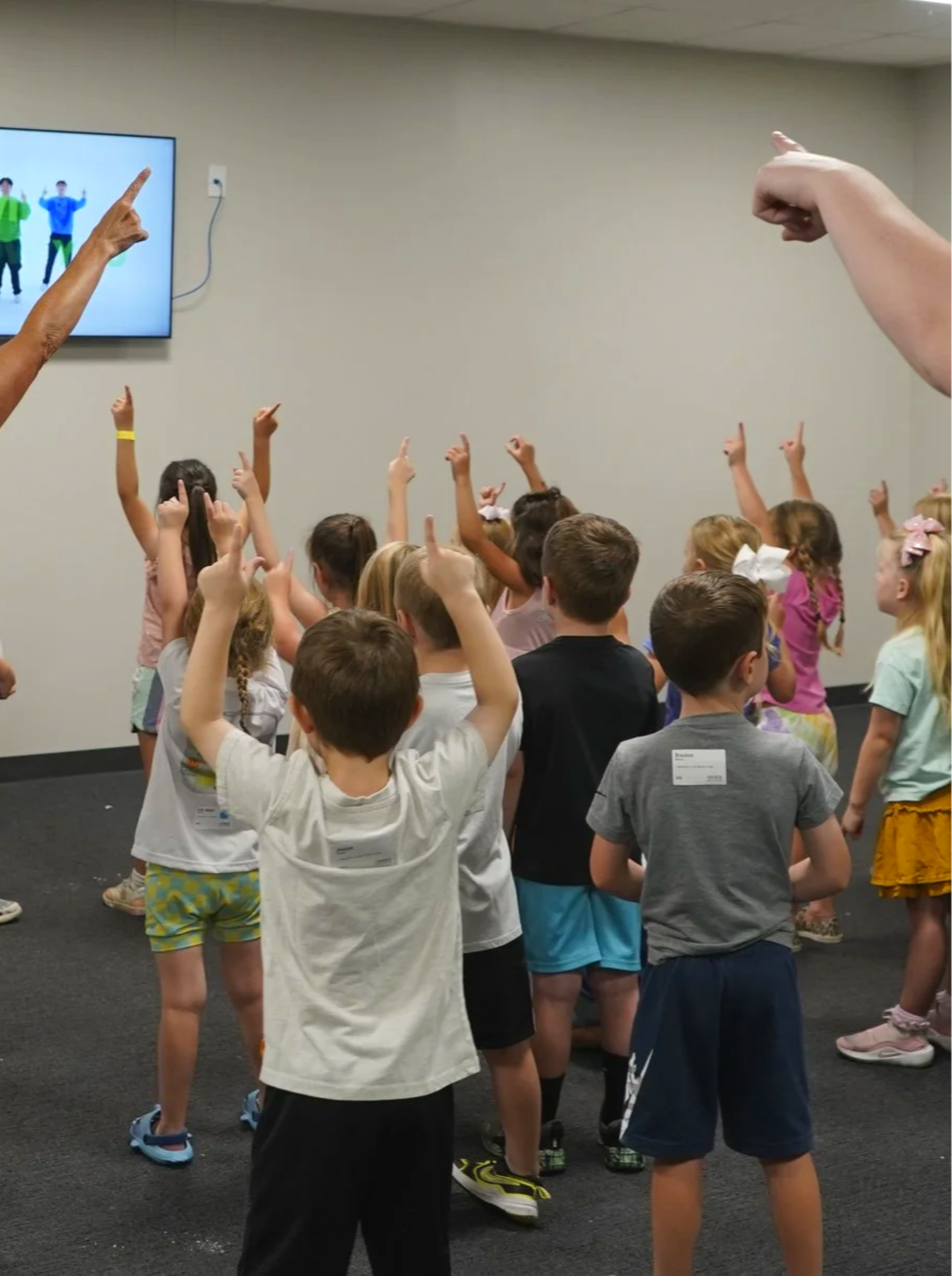 A group of children in a room with arms raised and pointing at a screen on the wall, participating in an activity or game.