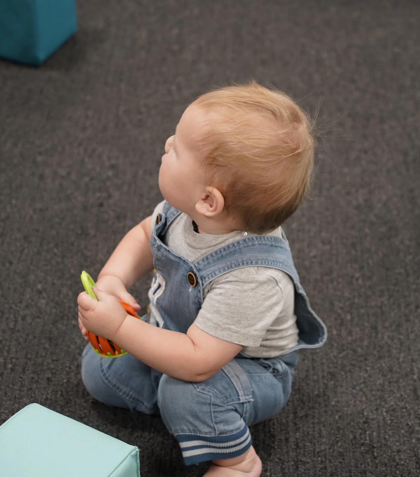 A young child with red hair sitting on the floor, holding a toy with orange and green colors, wearing a gray T-shirt and denim overalls.