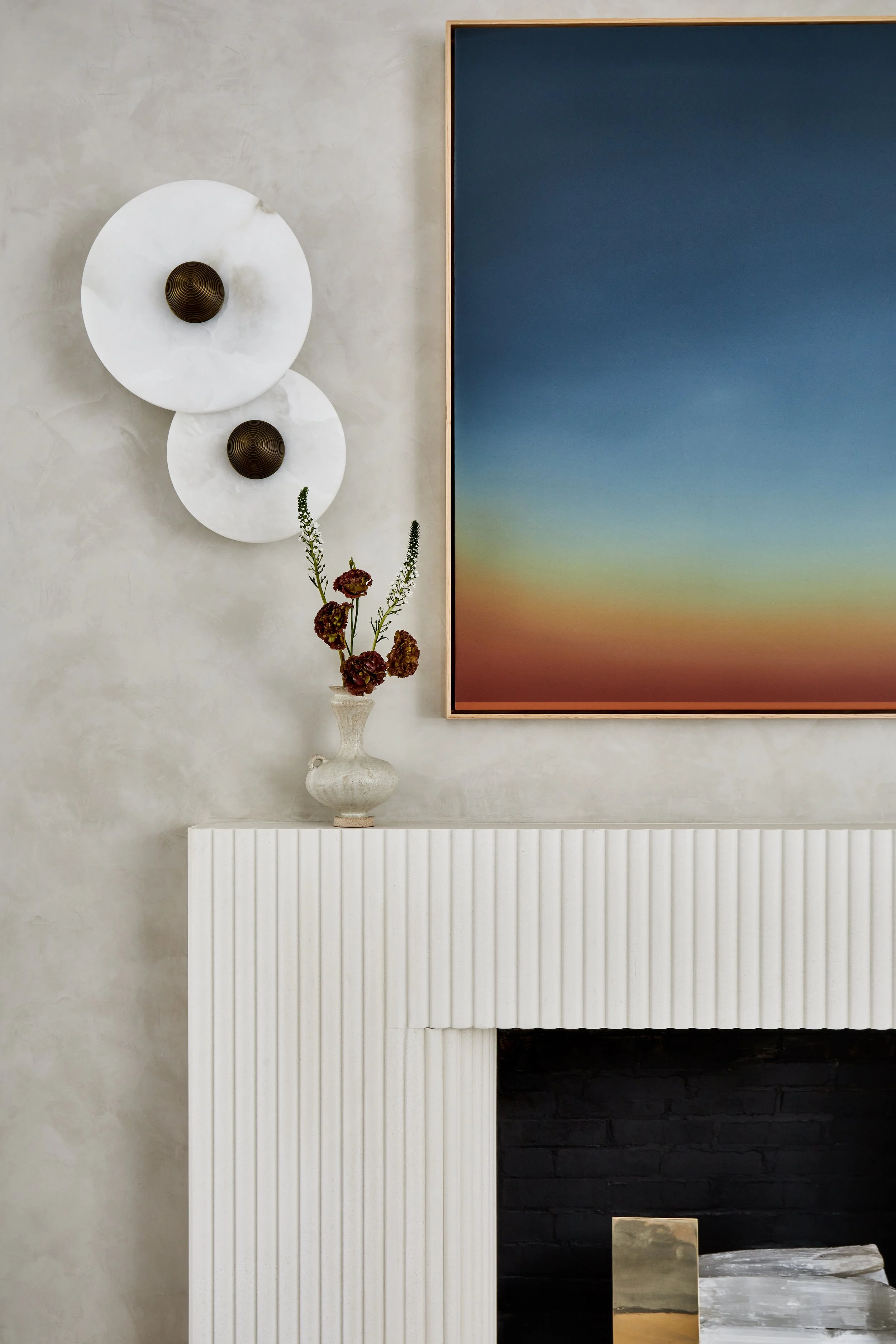 Decorative wall with two white circular wall sconces with bronze centers, a large framed gradient landscape photograph, a white vase with dried flowers, and part of a white fireplace mantel.