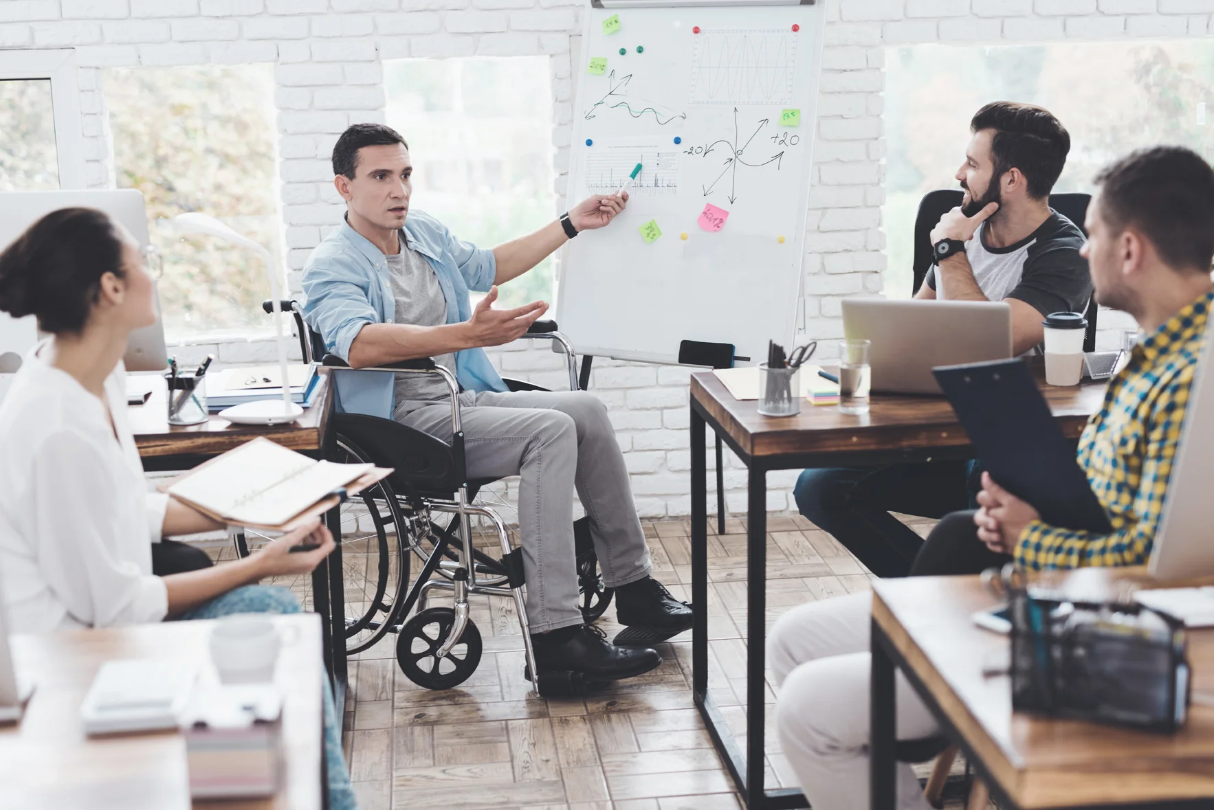 Man in a wheelchair pointing to a whiteboard and discussing graphic drawn on it with colleagues.