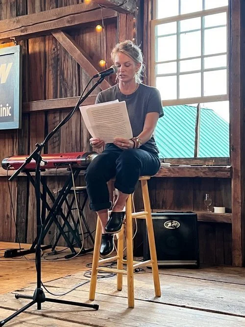 Woman reading a script on a stool at a microphone in a rustic wooden room with a keyboard, speaker, and window.