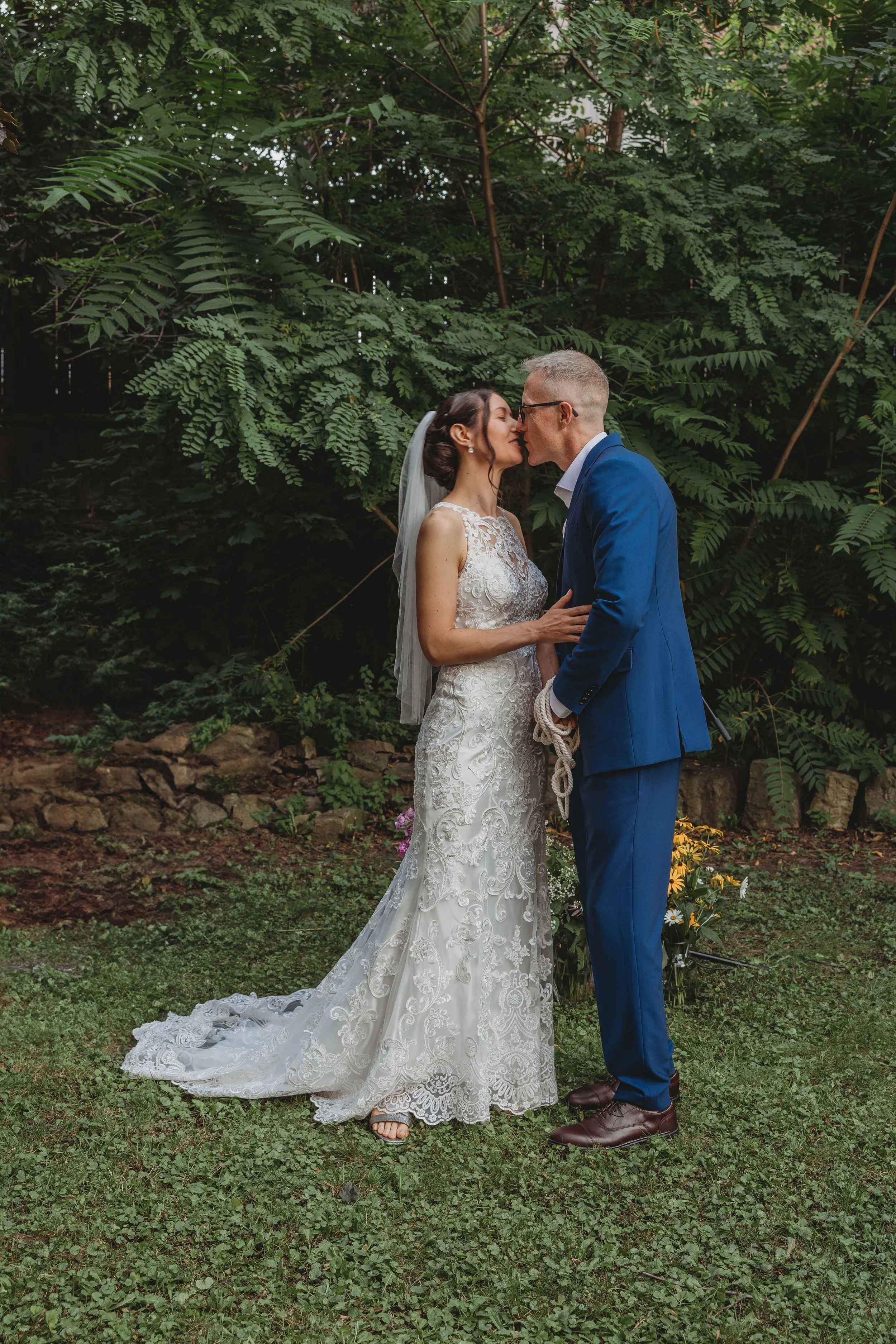 A bride and groom sharing a kiss outdoors, with the bride in a lace wedding gown and veil, and the groom in a blue suit, surrounded by green foliage.