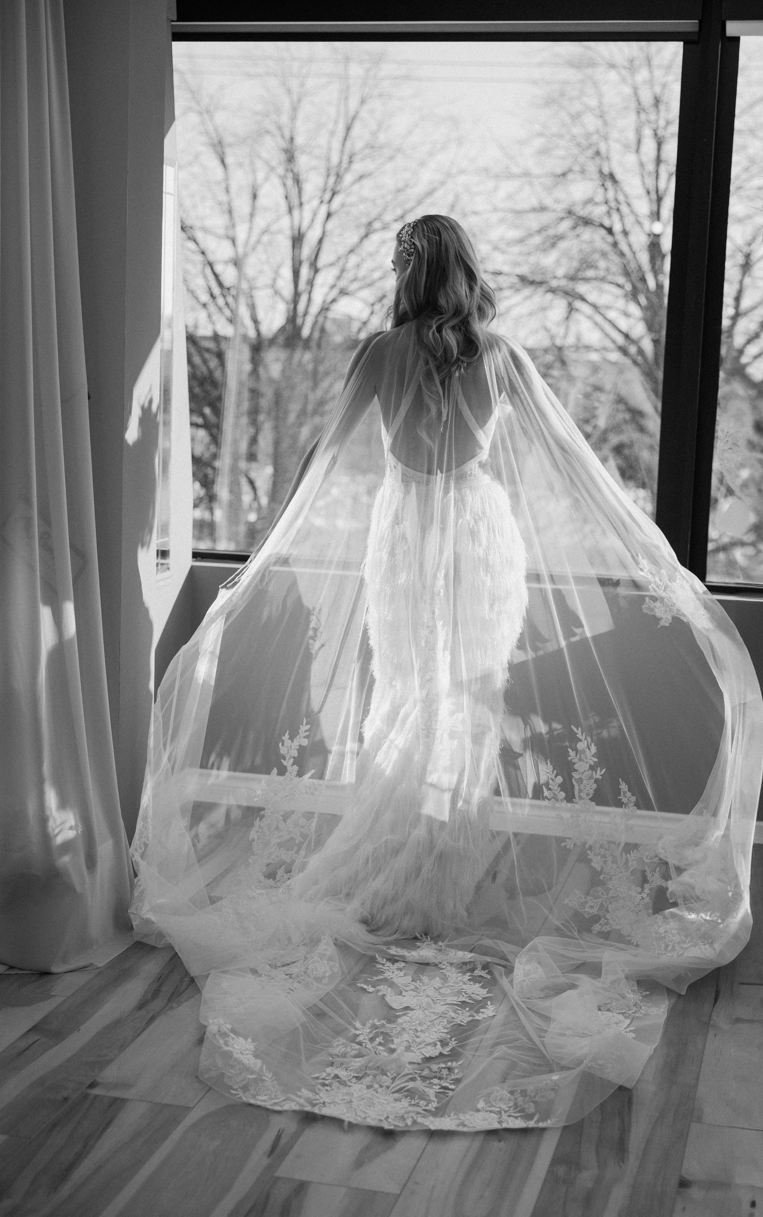A woman in a wedding dress standing by a large window, looking outside, with a long, flowing veil with lace details.