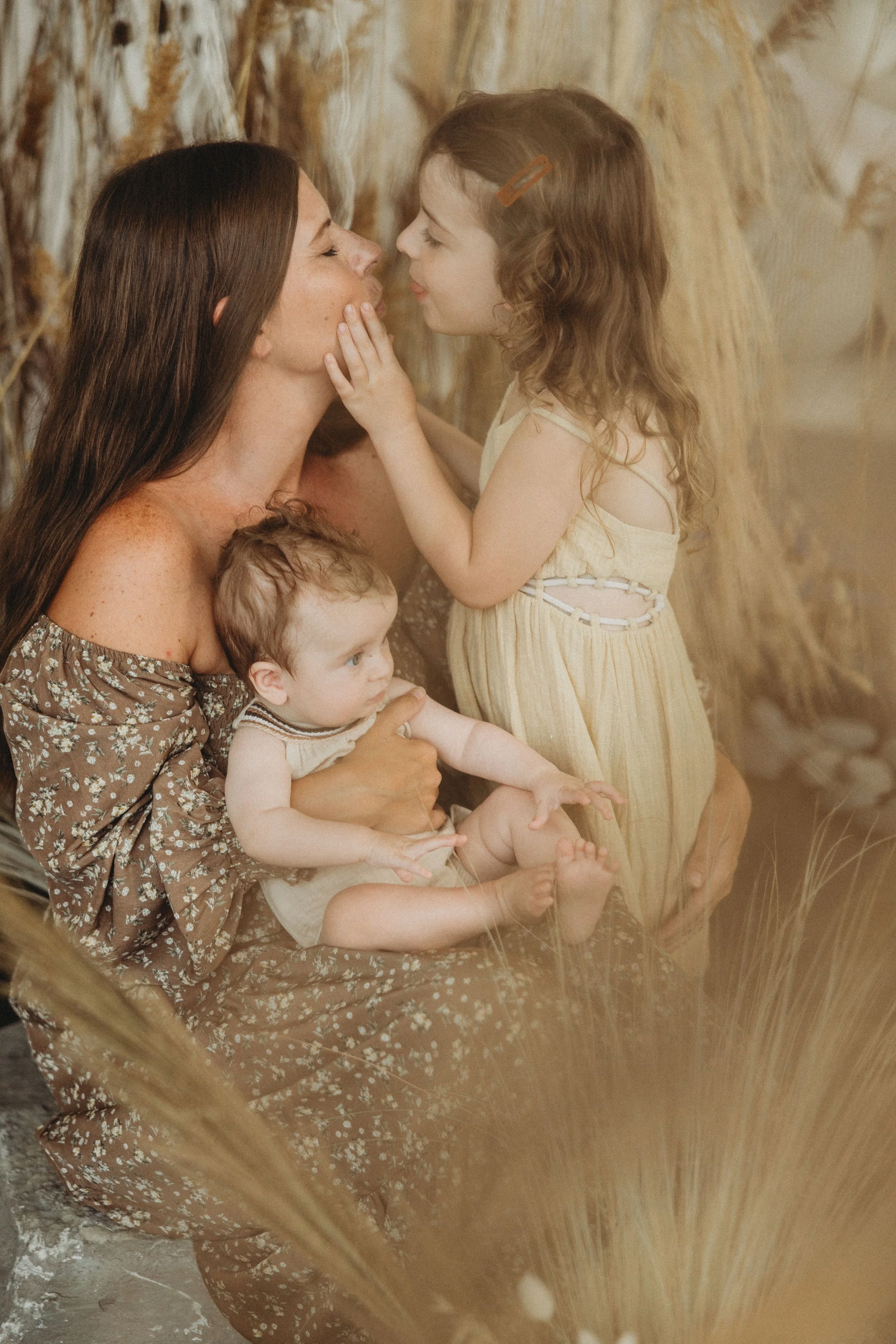 A woman with two young girls in a nature setting with dried grasses. One girl is touching the woman's face while the woman gently holds the other girl on her lap.