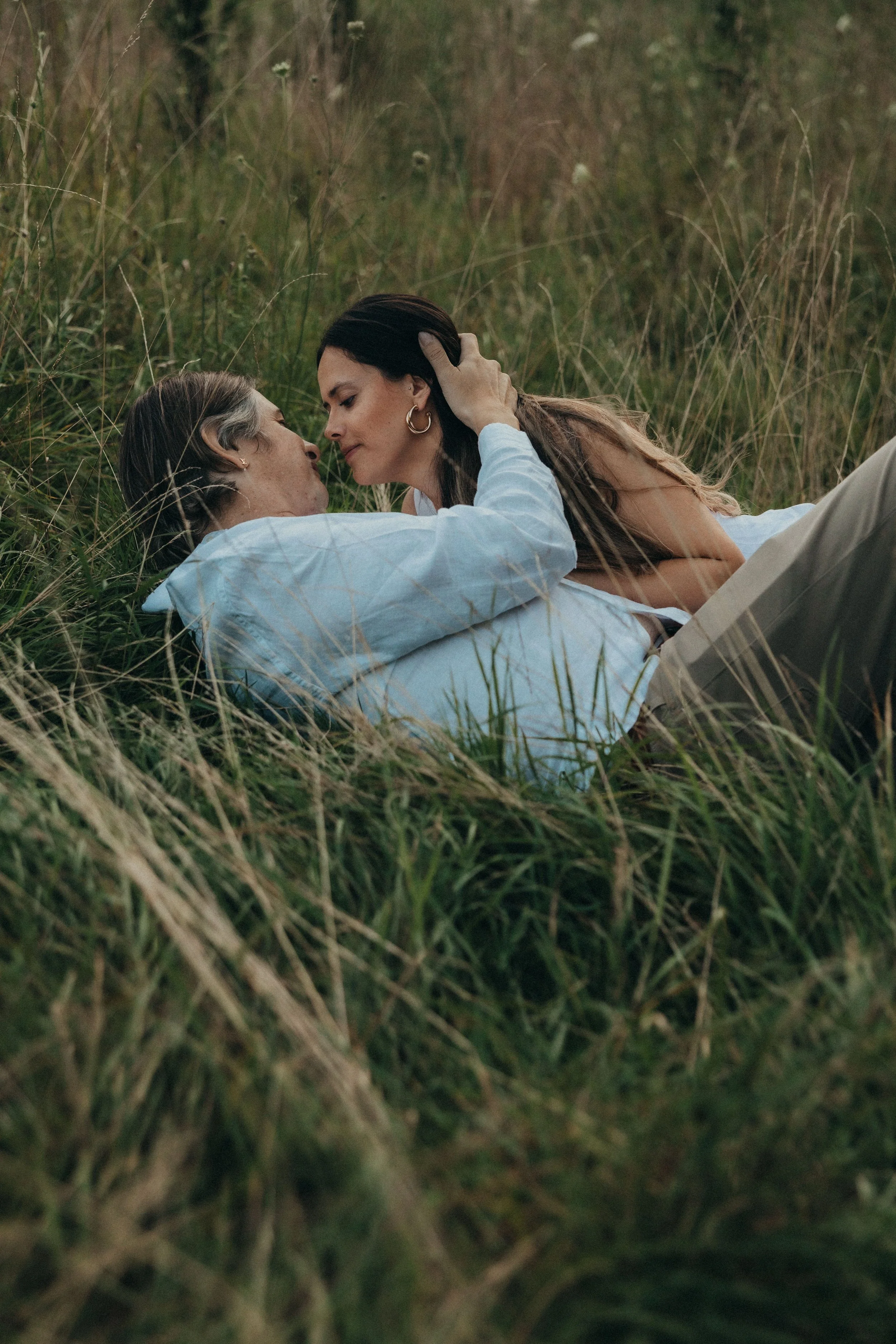 A man and woman lying on their sides in a grassy field, close and touching foreheads, sharing an intimate moment.