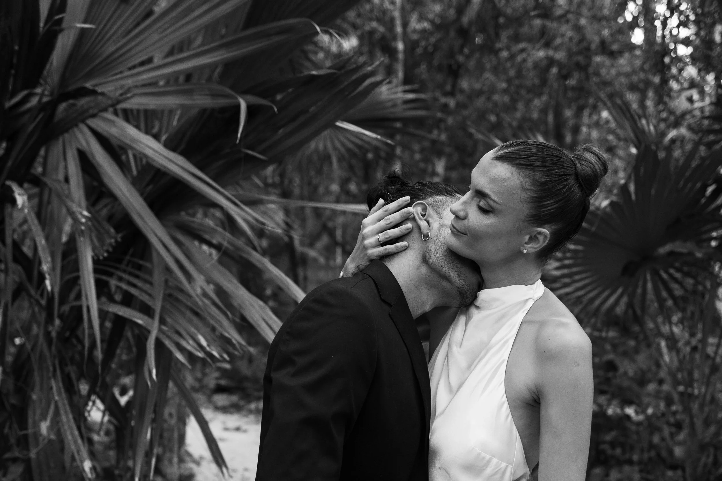 A woman in a white dress gently embraces a man in a dark suit, holding his head in a tender gesture in a lush outdoor setting with large tropical leaves.