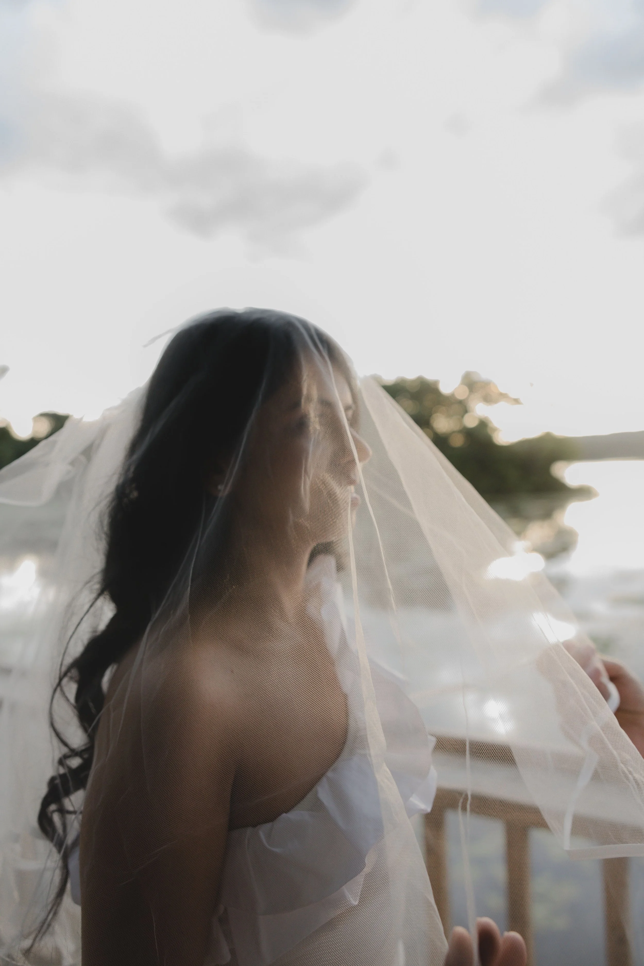 A bride and groom sharing a kiss outdoors, with the bride in a lace wedding gown and veil, and the groom in a blue suit, surrounded by green foliage.