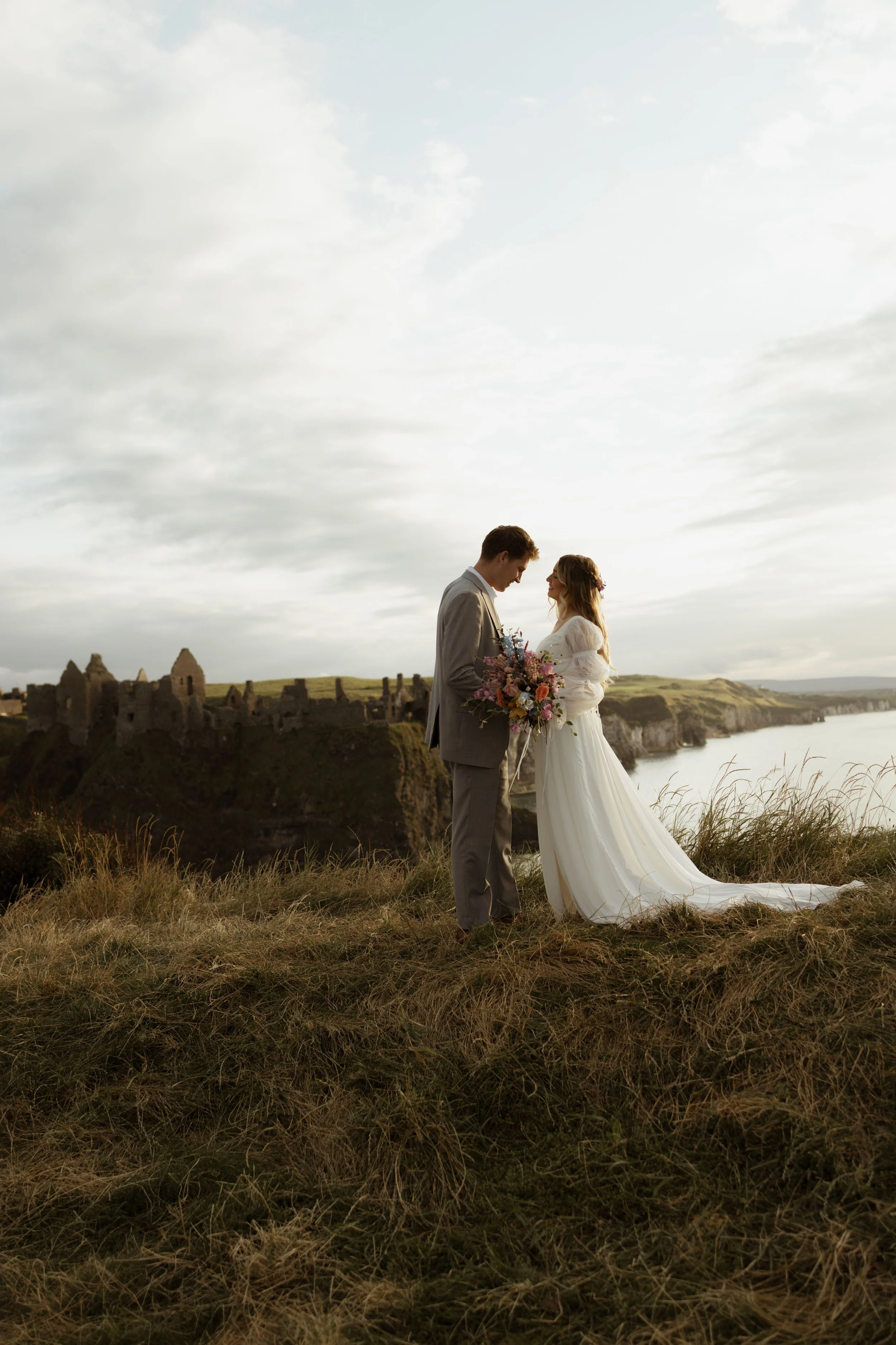 A woman in a wedding dress standing by a large window, looking outside, with a long, flowing veil with lace details.