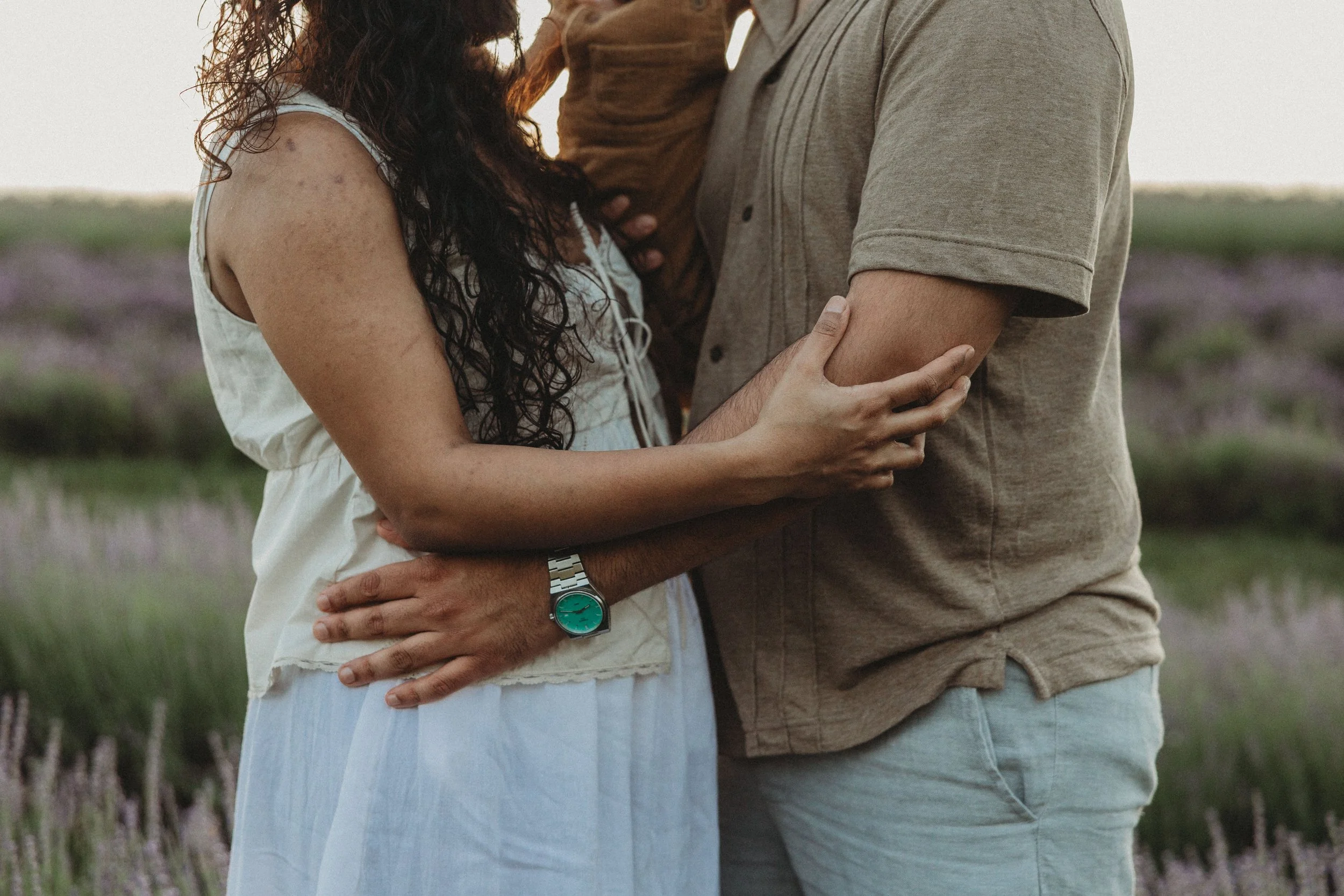 A woman in a white dress gently embraces a man in a dark suit, holding his head in a tender gesture in a lush outdoor setting with large tropical leaves.