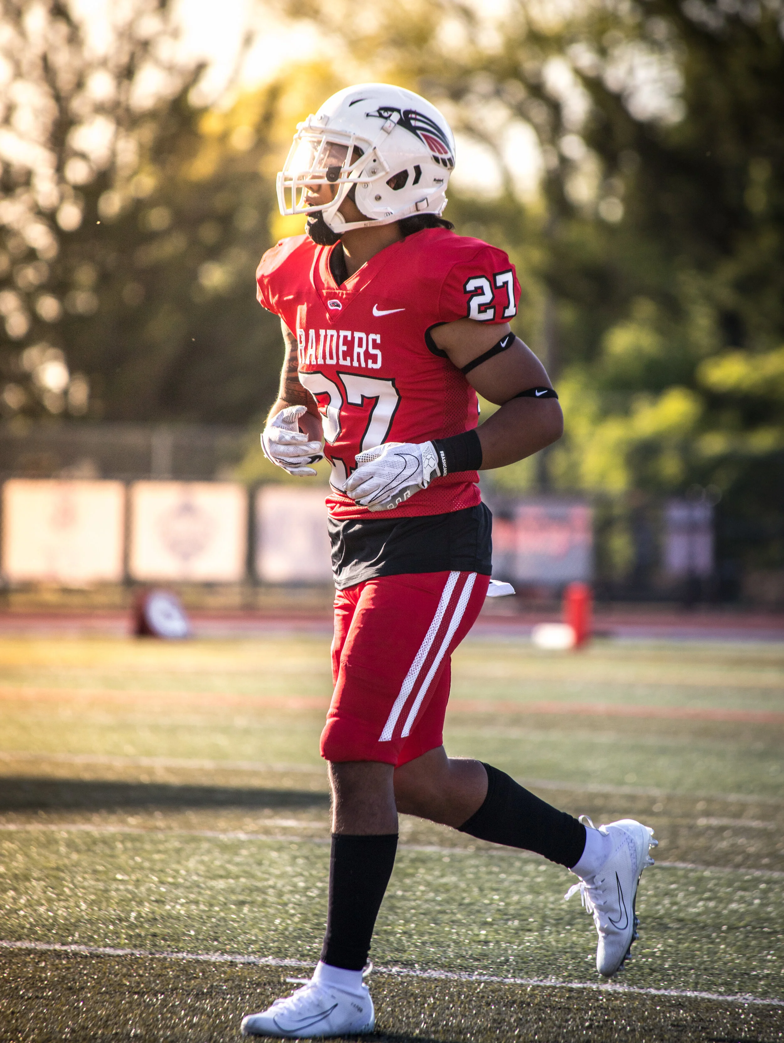 A football player in a red uniform, number 27, standing on a football field at sunset, wearing a white helmet with a logo, black gloves, and white cleats.