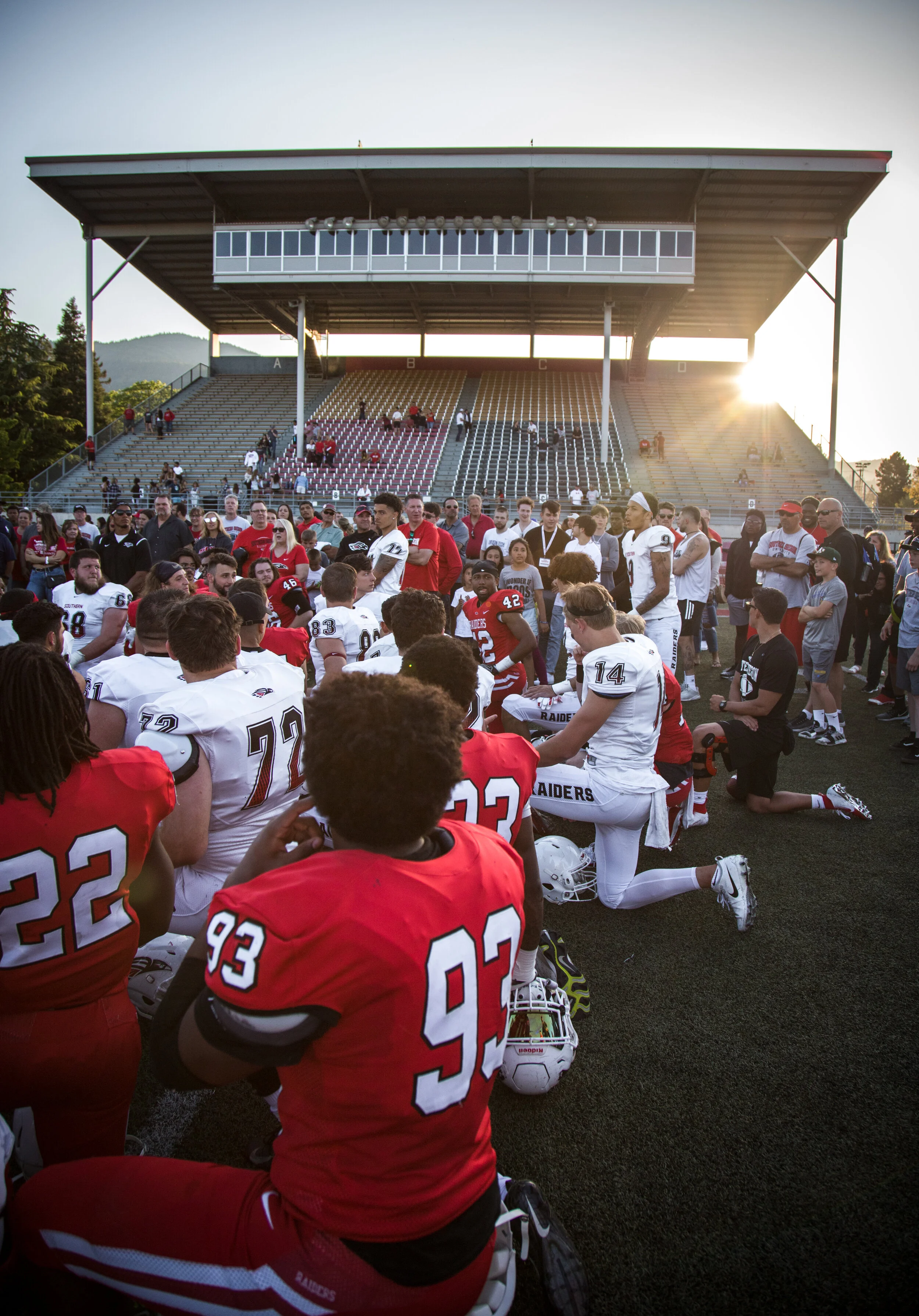 Football players in red and white uniforms kneeling and standing on a football field during a game, with a large crowd and bleachers in the background at sunset.