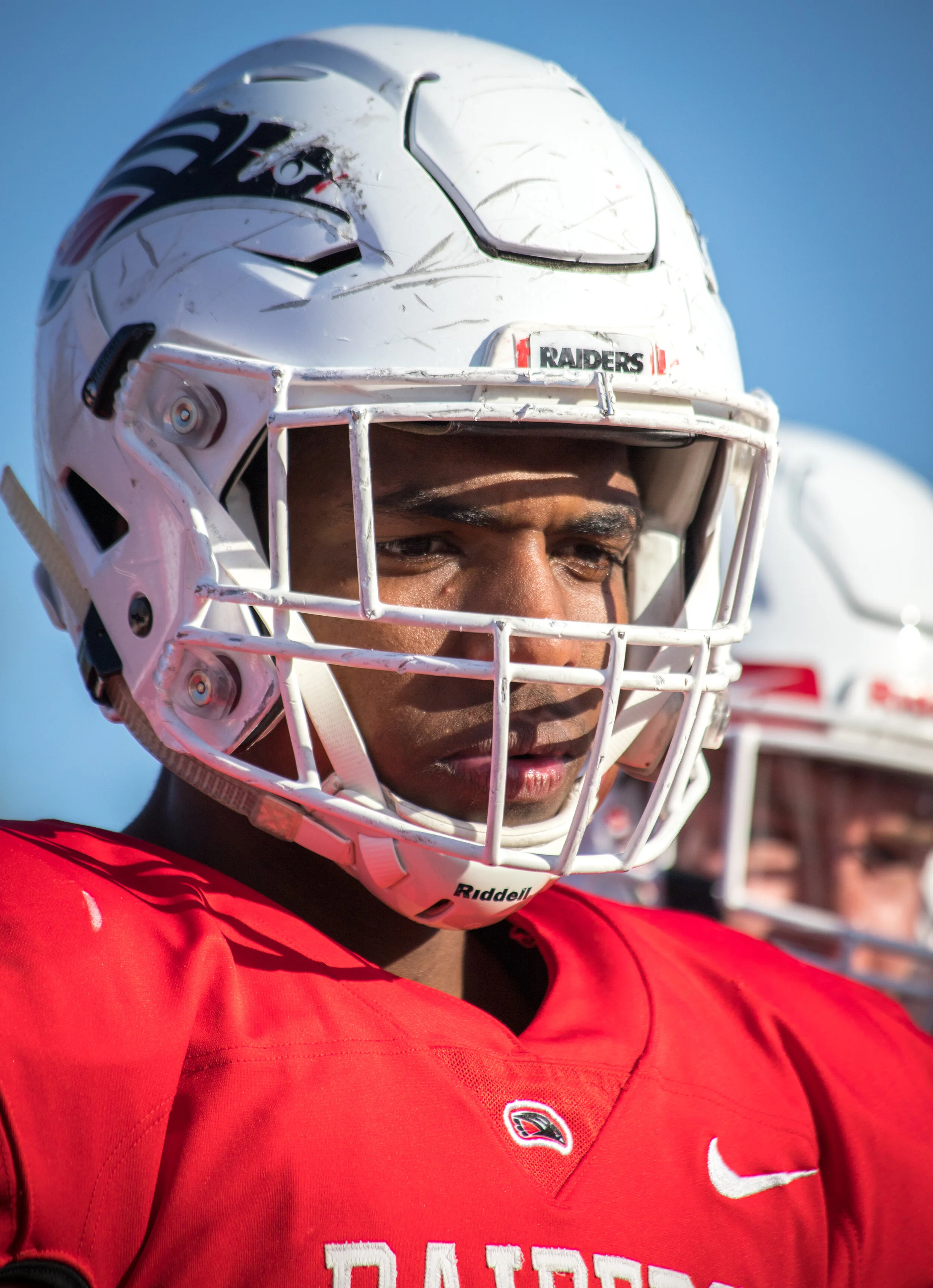 Close-up of a football player wearing a white helmet with a faceguard, and a red jersey with the word "Raiders" and a Nike logo. The background shows a clear blue sky.