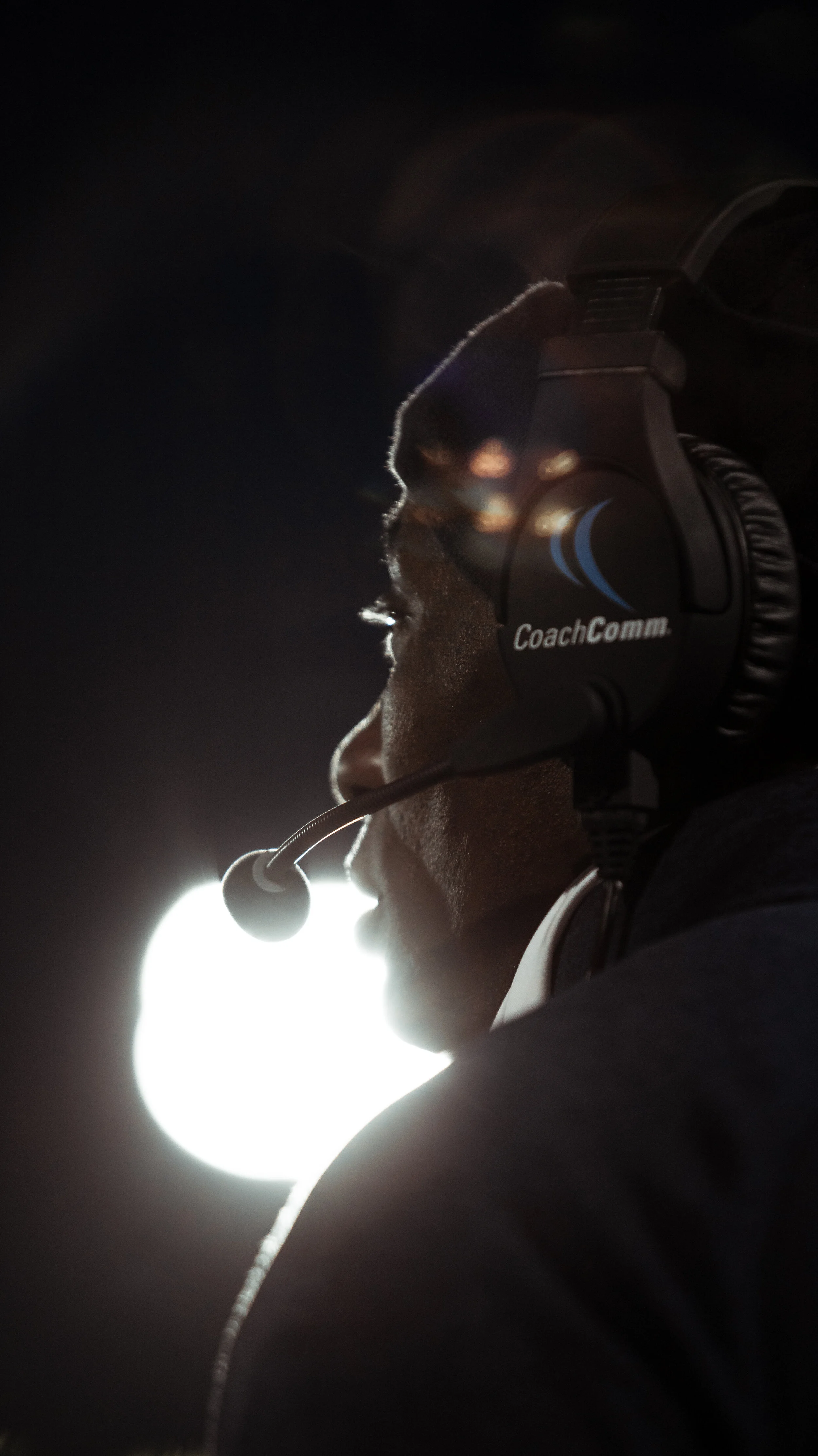 Close-up of a person wearing a CoachComm headset with a microphone, side profile, dark background with bright light in the distance.